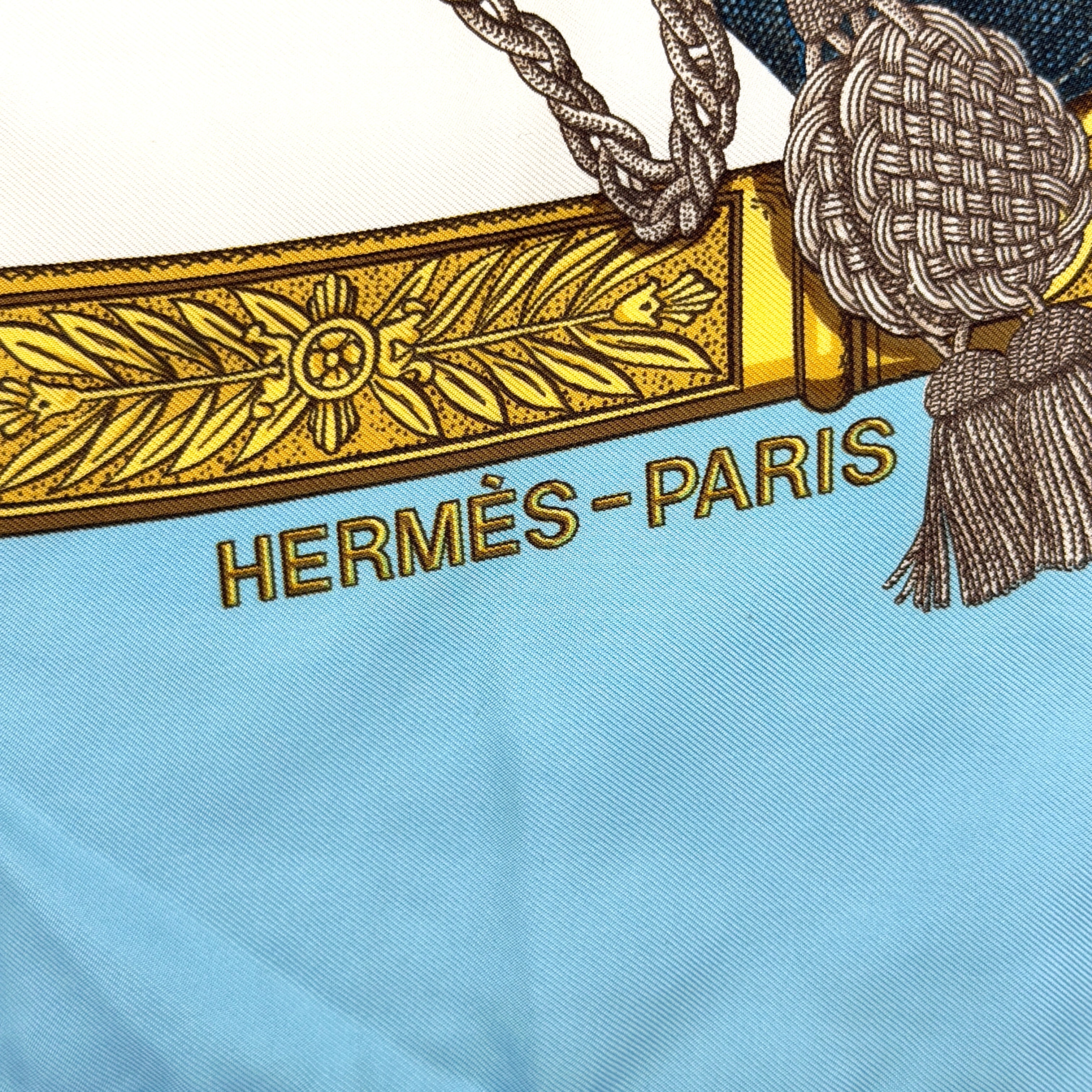 Hermès Grand Uniforme silk scarf with intricate gold and blue Napoleonic-inspired motifs framed on a soft blue and ivory background.