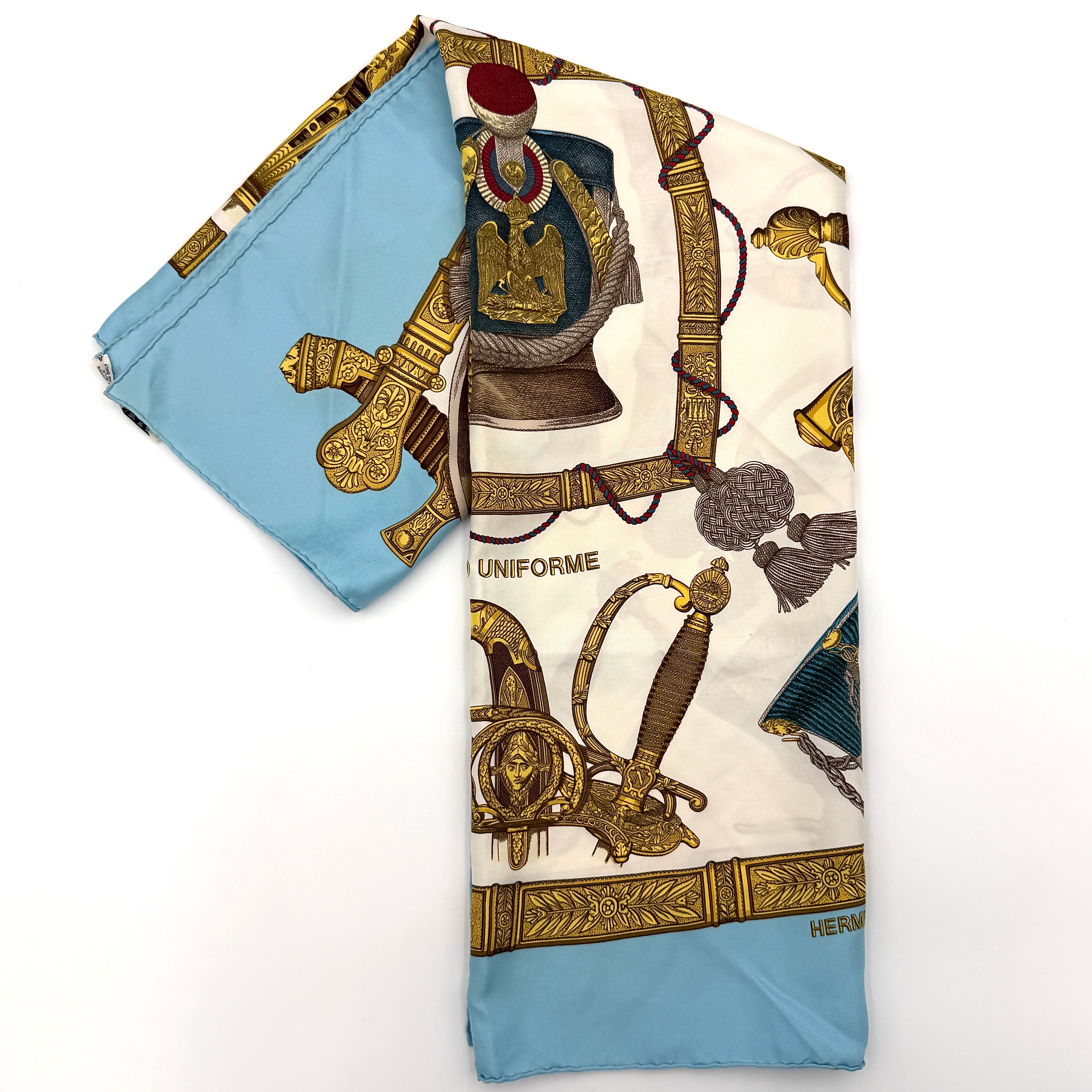 Hermès Grand Uniforme silk scarf with intricate gold and blue Napoleonic-inspired motifs framed on a soft blue and ivory background.