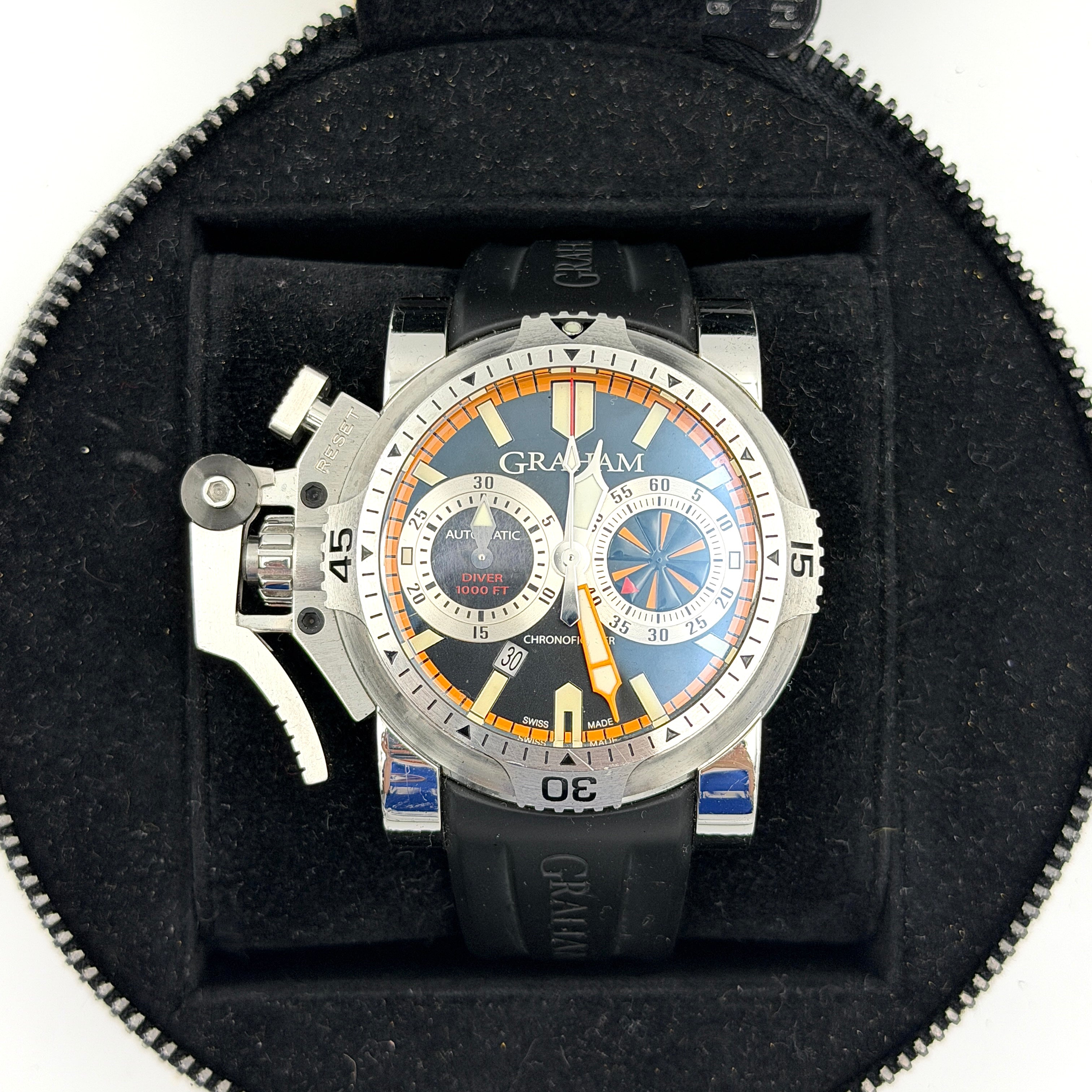 The Graham Chronofighter Oversize Diver 2OVES.B15A watch displayed in its signature round Graham London presentation case, showcasing its bold black dial, prominent chronograph trigger, and black rubber strap.