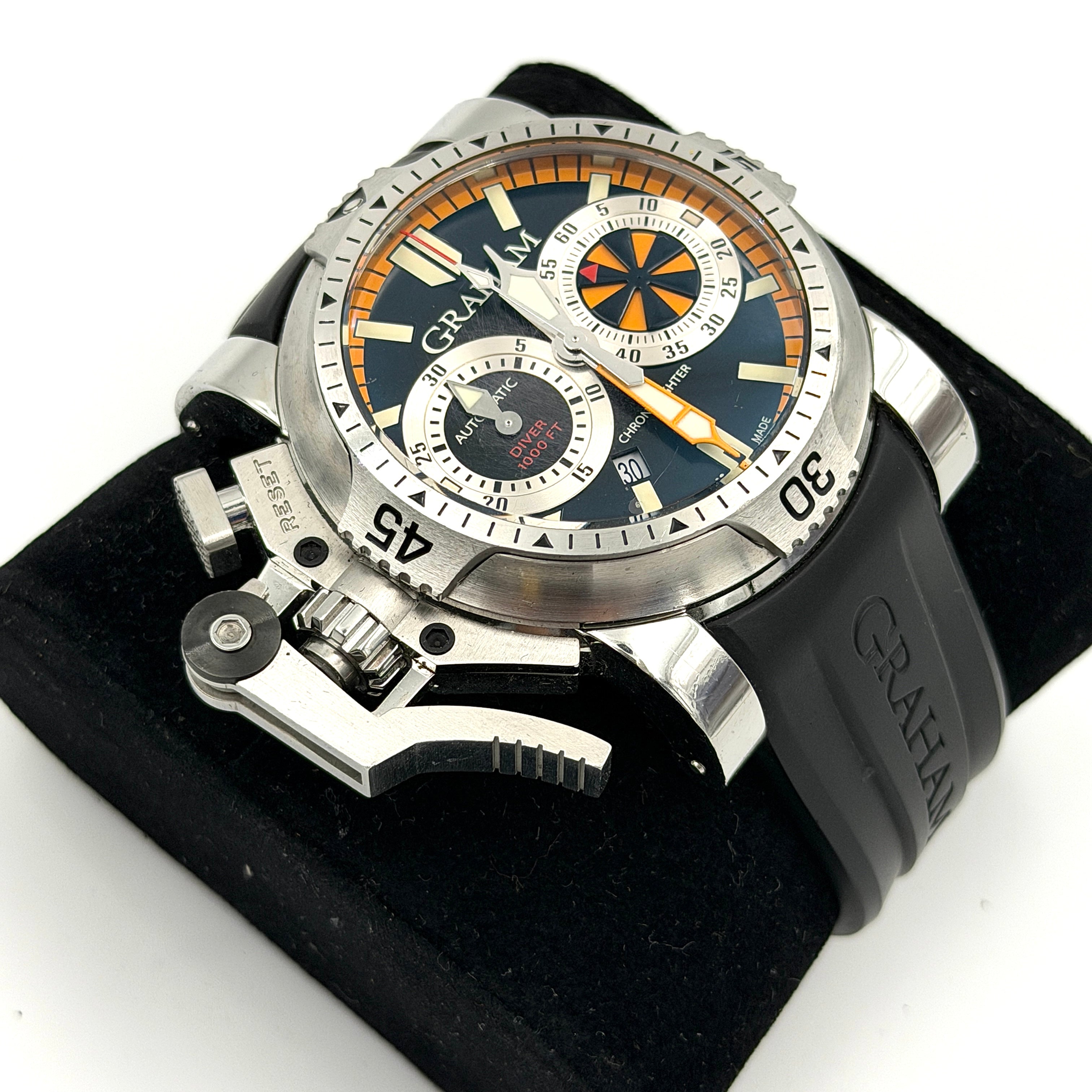 The Graham Chronofighter Oversize Diver 2OVES.B15A watch displayed in its signature round Graham London presentation case, showcasing its bold black dial, prominent chronograph trigger, and black rubber strap.
