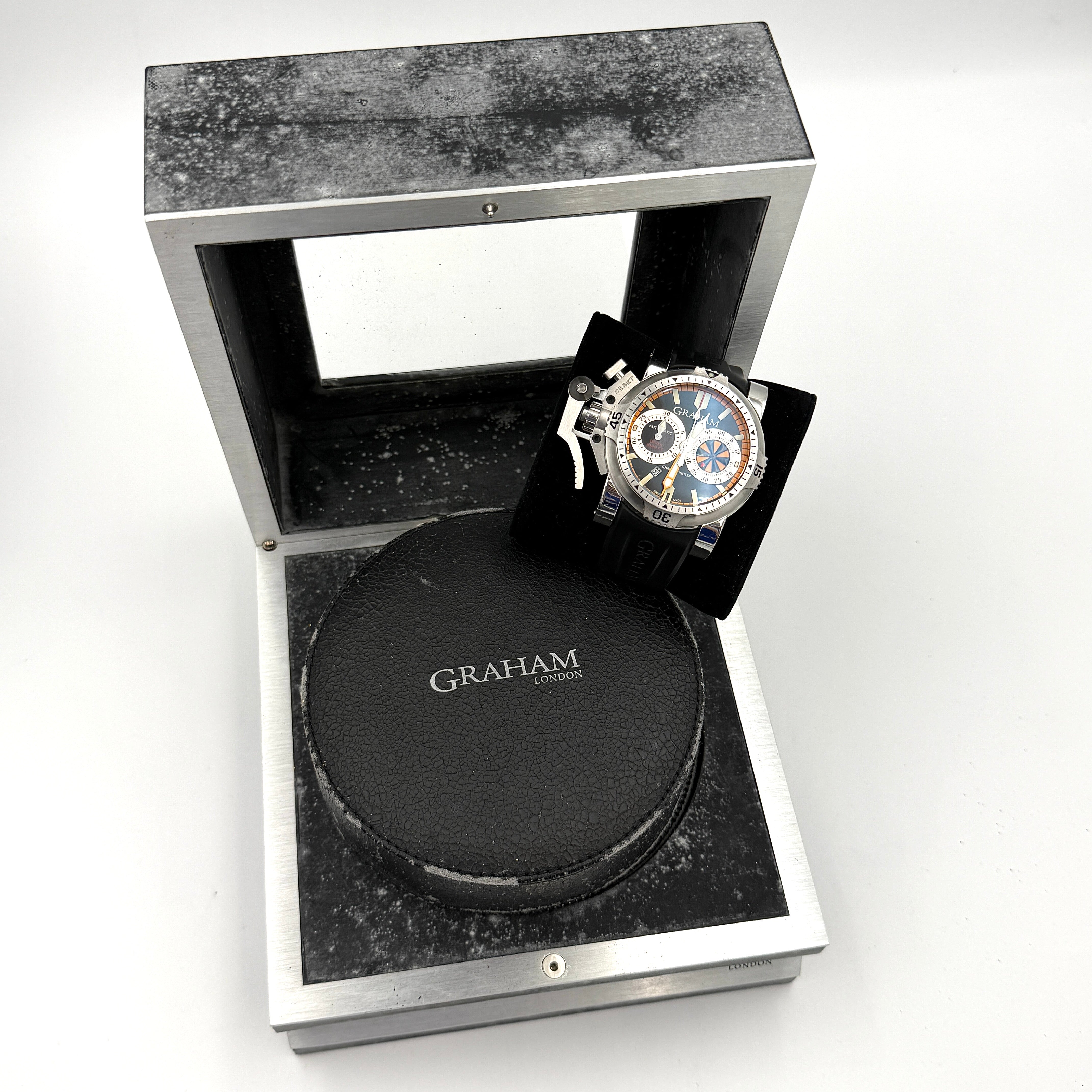 The Graham Chronofighter Oversize Diver 2OVES.B15A watch displayed in its signature round Graham London presentation case, showcasing its bold black dial, prominent chronograph trigger, and black rubber strap.