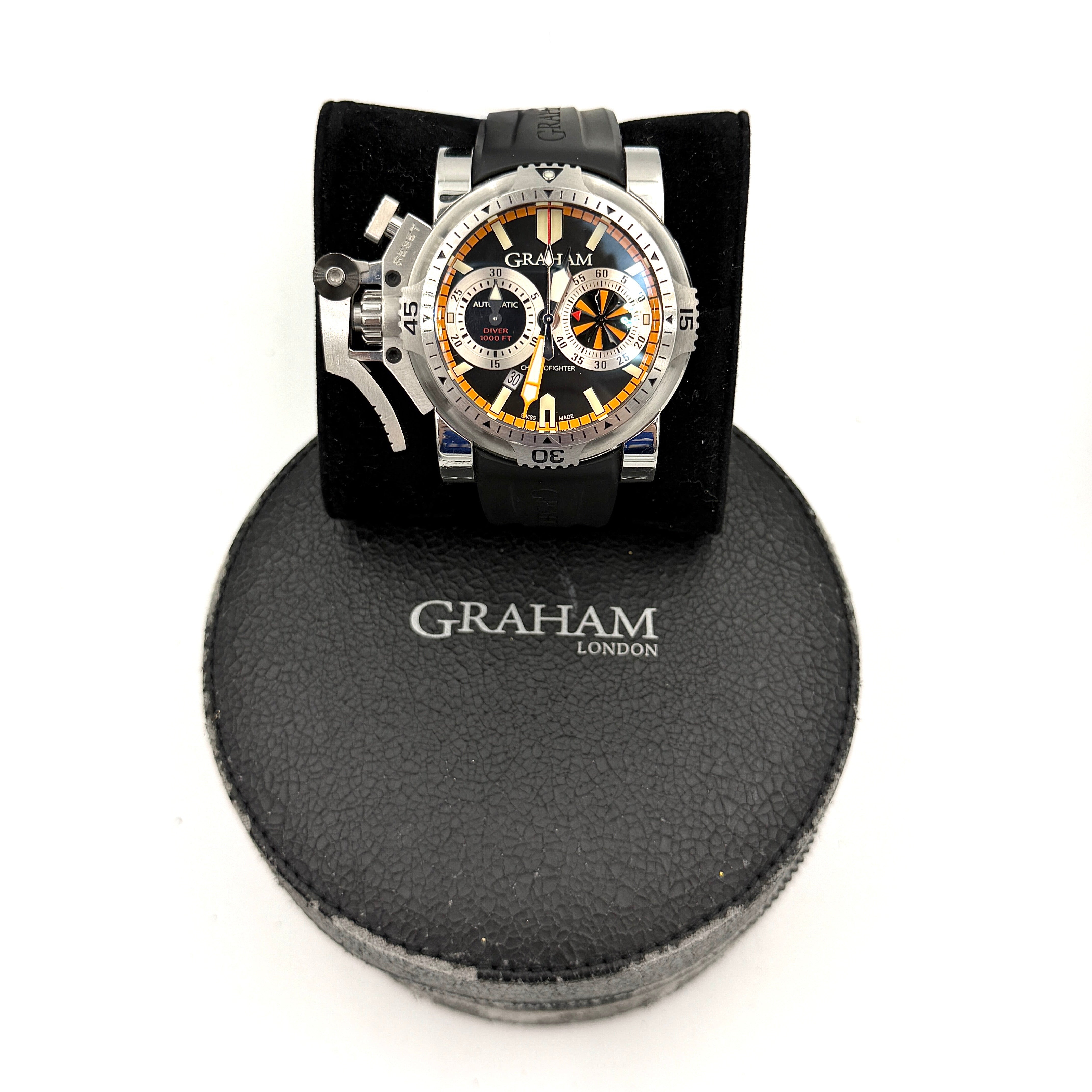 The Graham Chronofighter Oversize Diver 2OVES.B15A watch displayed in its signature round Graham London presentation case, showcasing its bold black dial, prominent chronograph trigger, and black rubber strap.