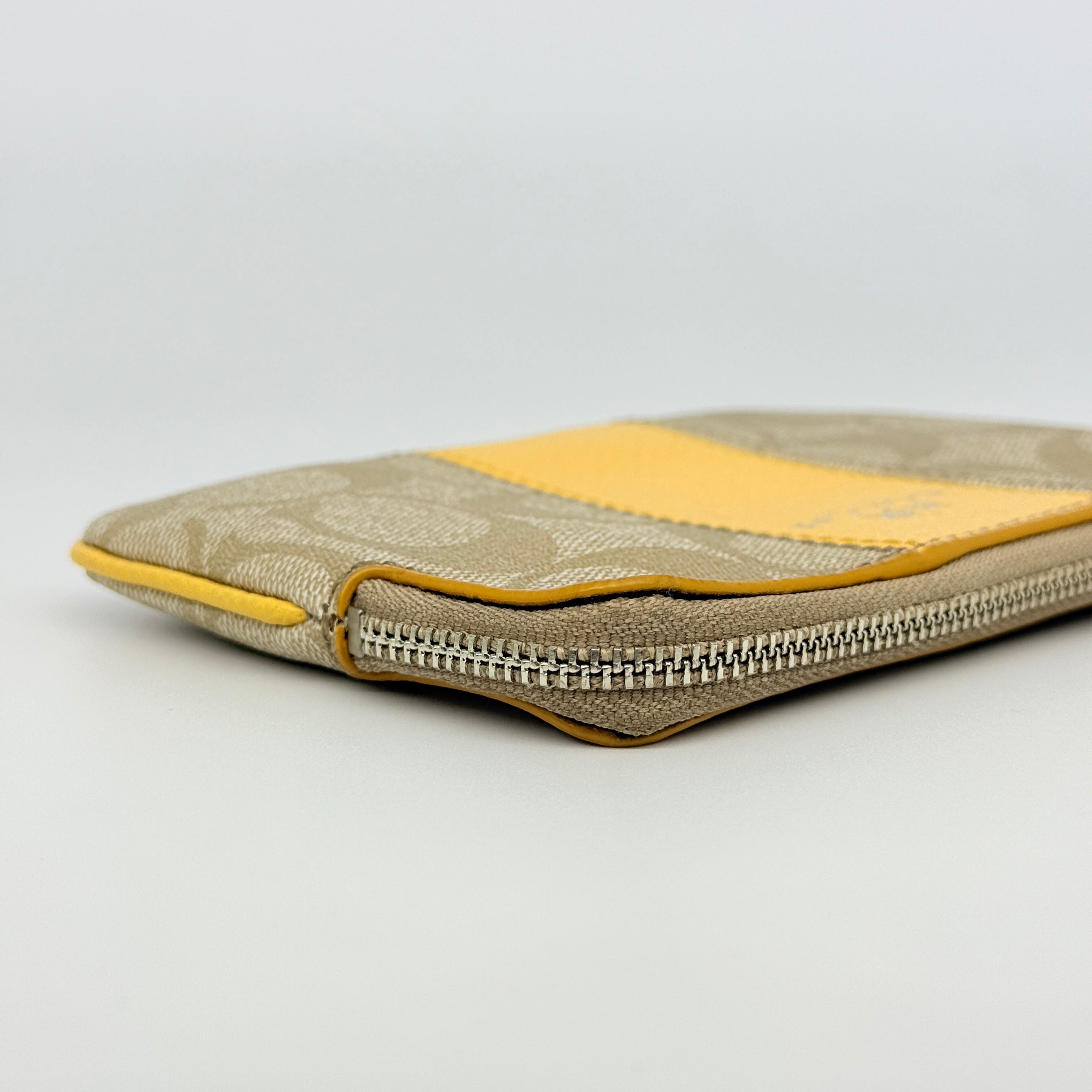 Coach Signature Stripe Corner Zip Wristlet in beige canvas with bold yellow leather stripe, silver hardware, and wrist strap displayed on a white background.