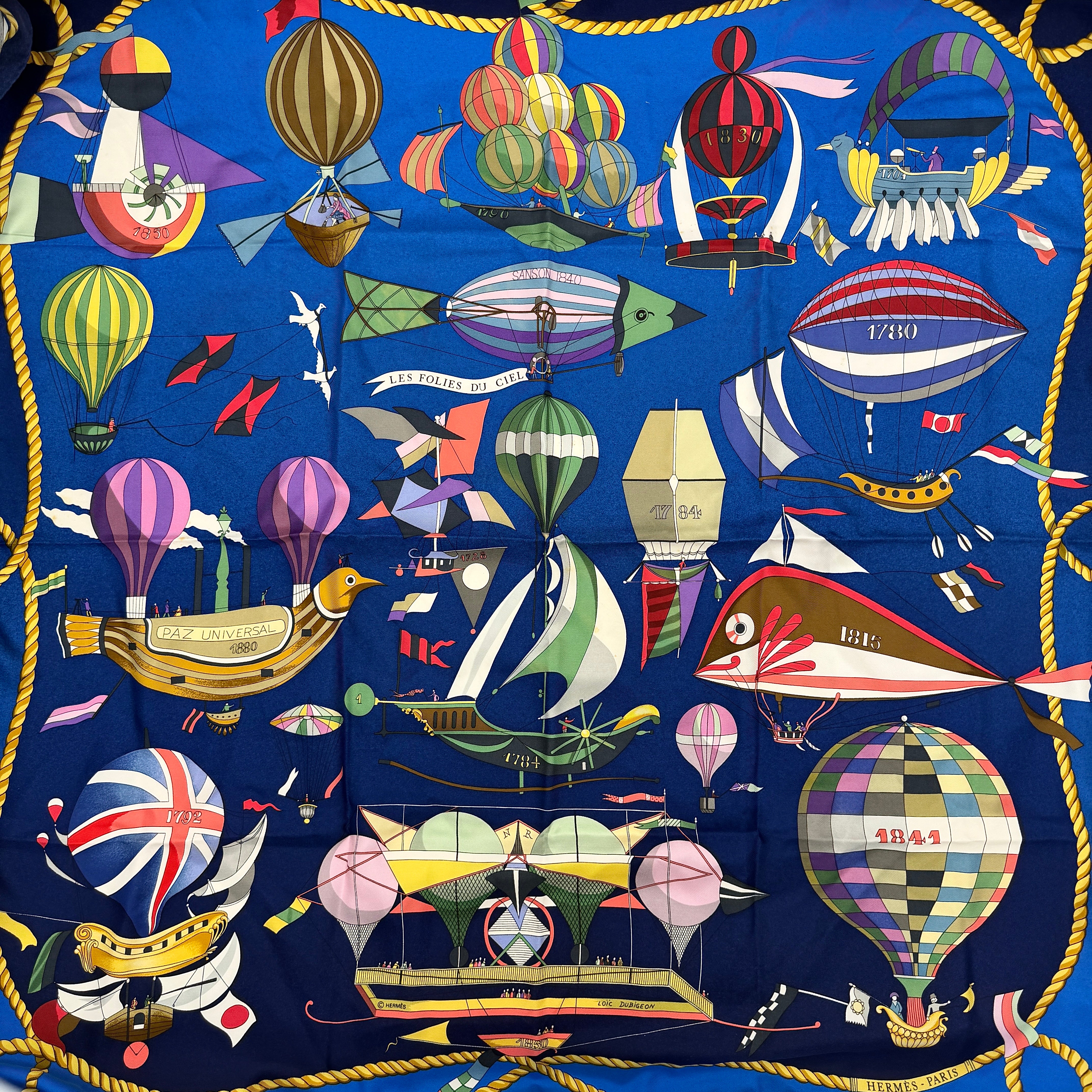 Hermès Les Folies du Ciel silk scarf in vivid multicolours with whimsical hot air balloons and golden rope border on a blue background.