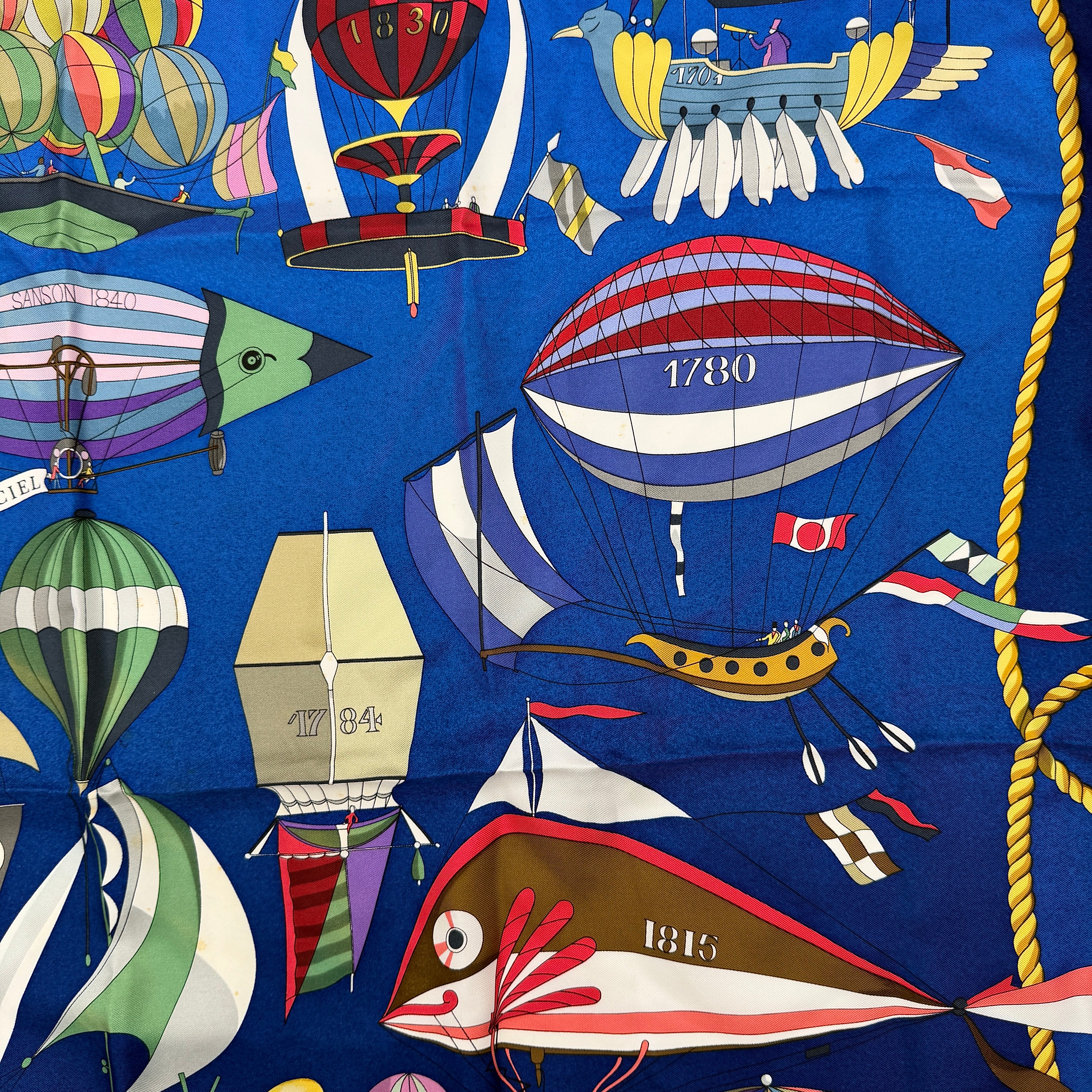 Hermès Les Folies du Ciel silk scarf in vivid multicolours with whimsical hot air balloons and golden rope border on a blue background.