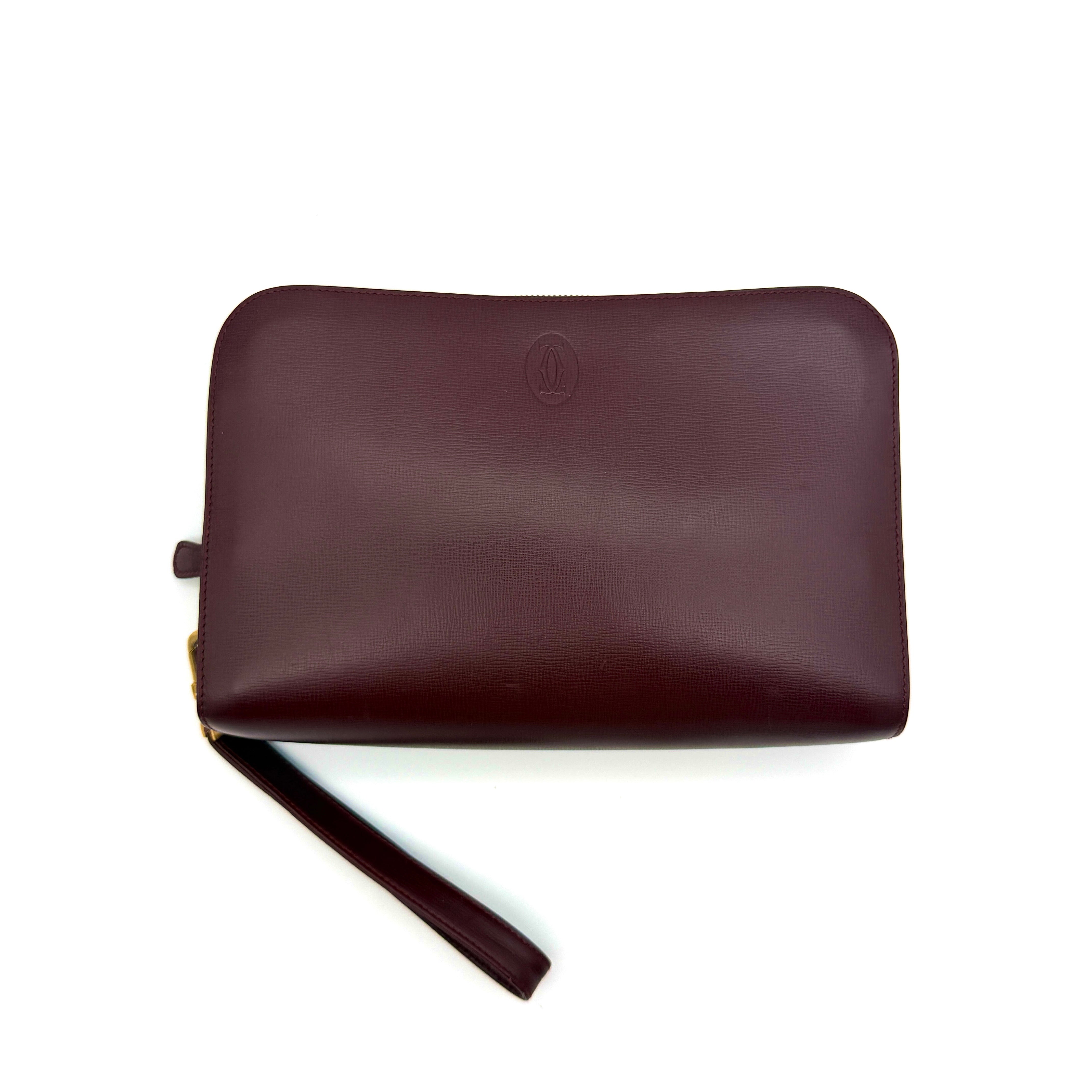 Cartier Must de Cartier clutch in burgundy leather with gold-tone zip and wrist strap, shown against a white background.