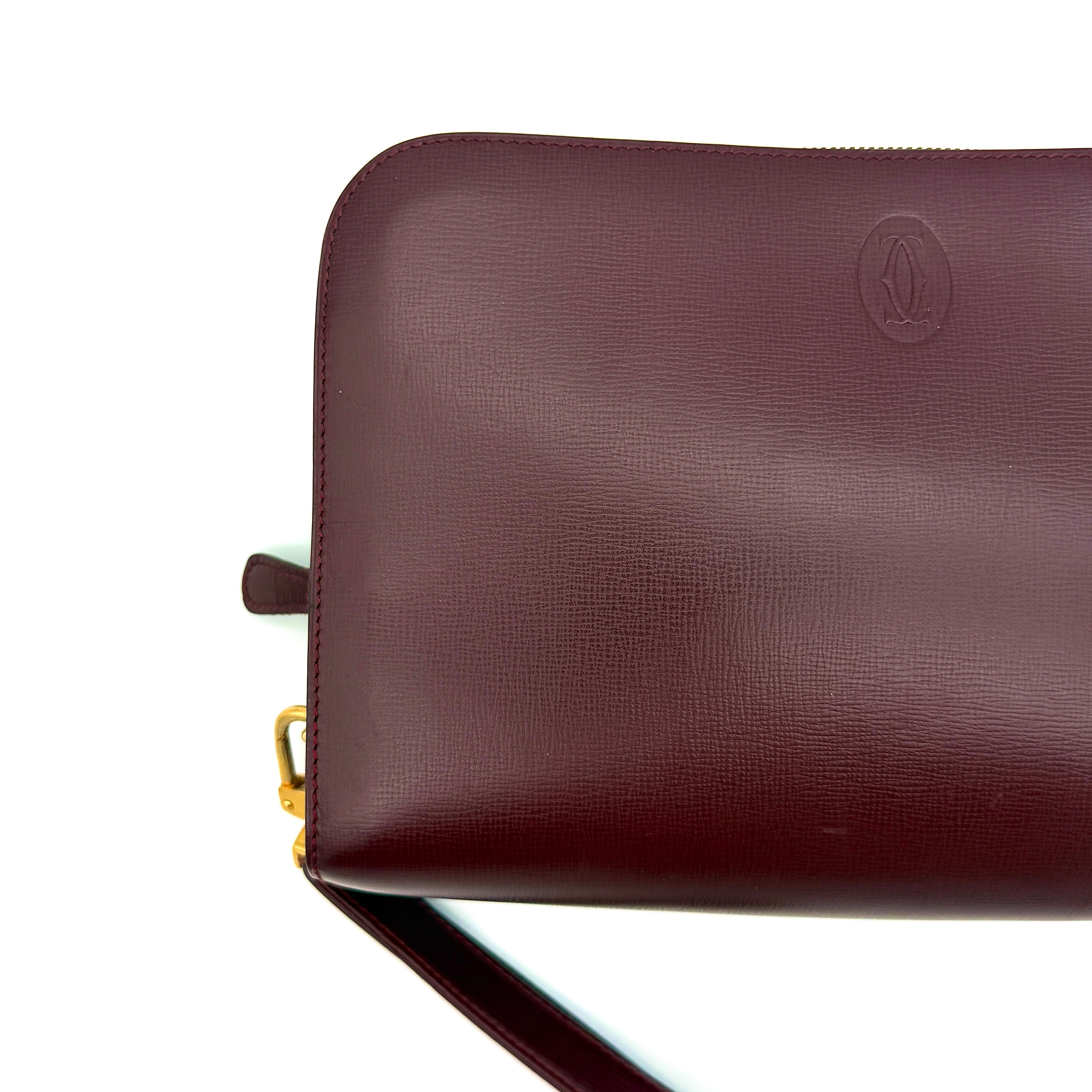 Cartier Must de Cartier clutch in burgundy leather with gold-tone zip and wrist strap, shown against a white background.