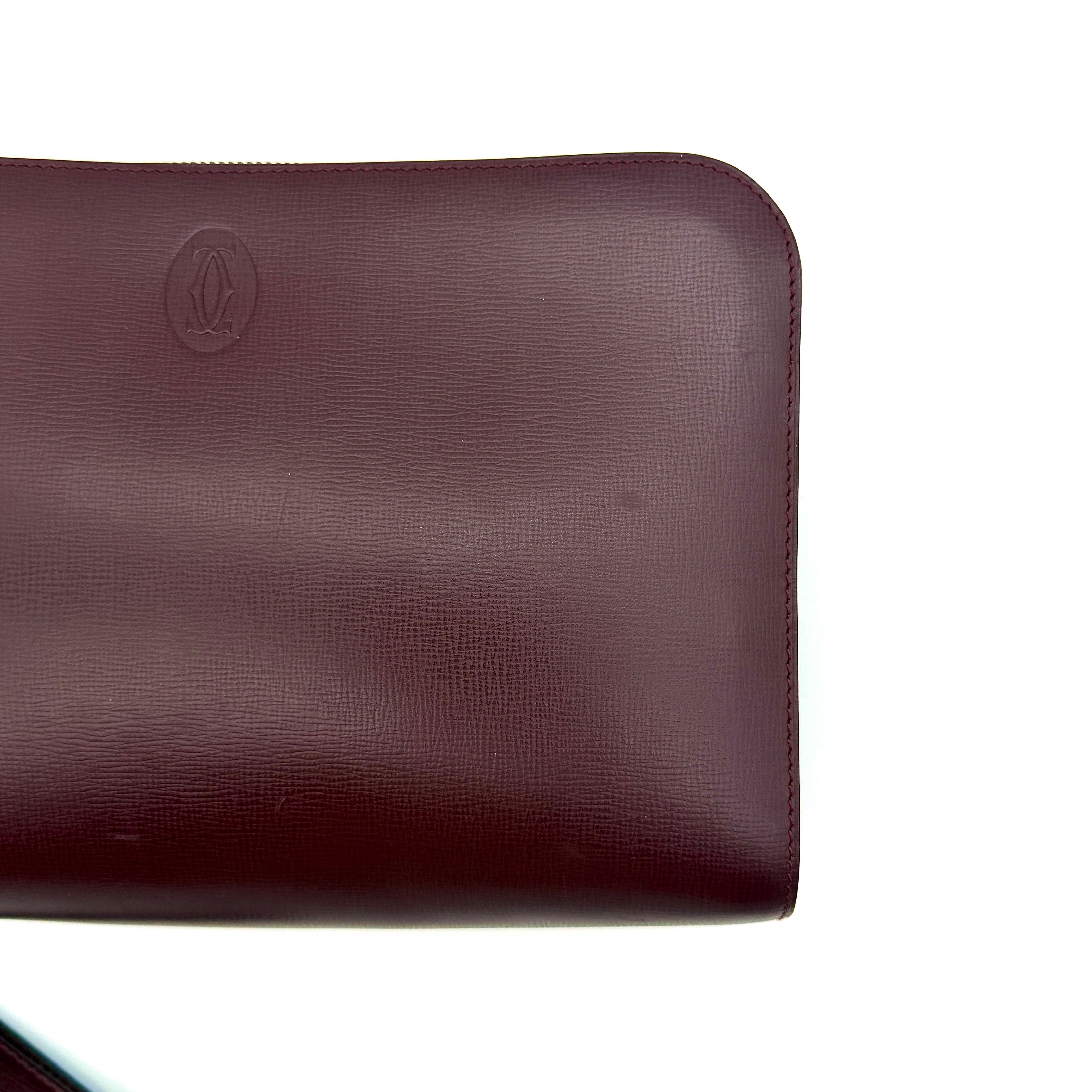 Cartier Must de Cartier clutch in burgundy leather with gold-tone zip and wrist strap, shown against a white background.