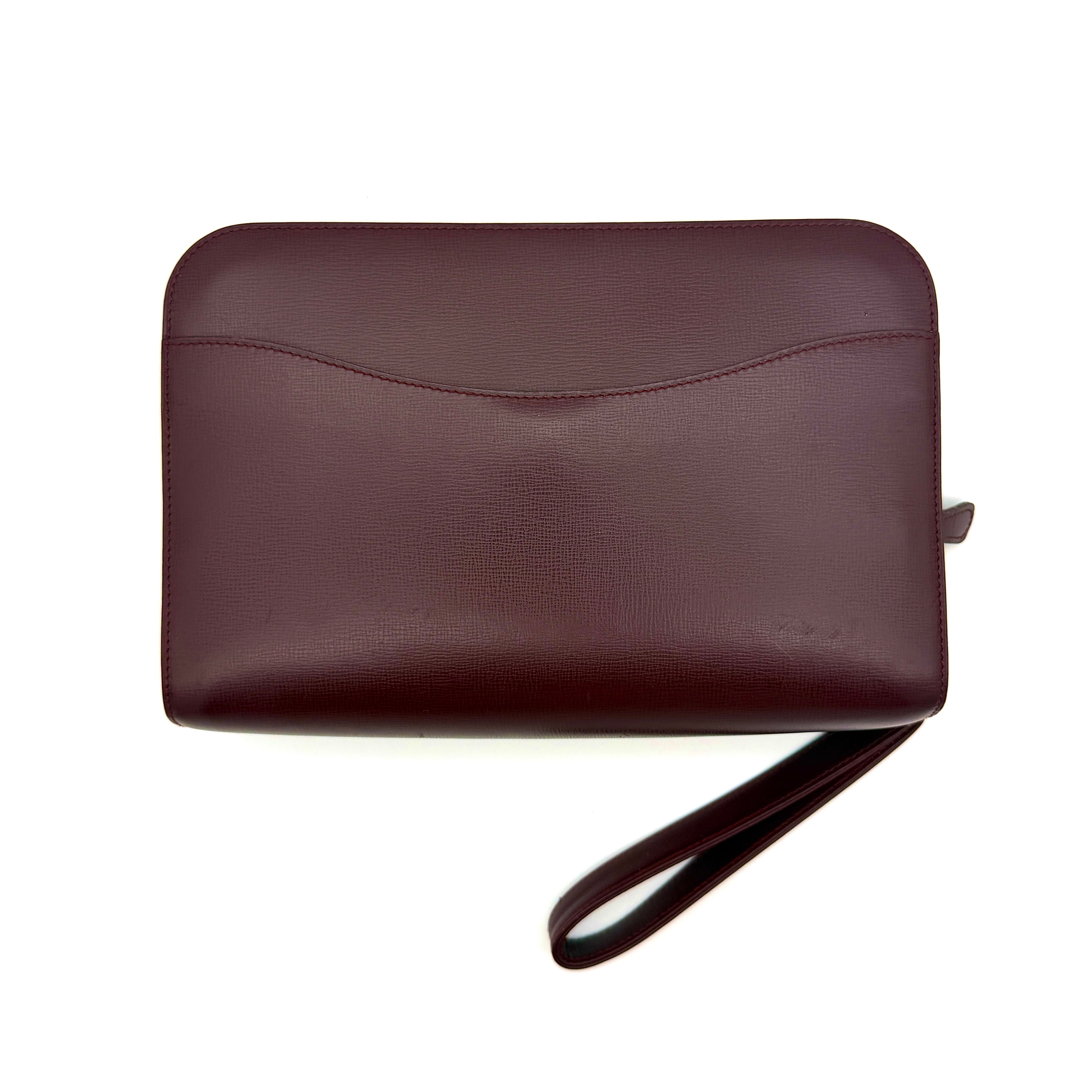 Cartier Must de Cartier clutch in burgundy leather with gold-tone zip and wrist strap, shown against a white background.