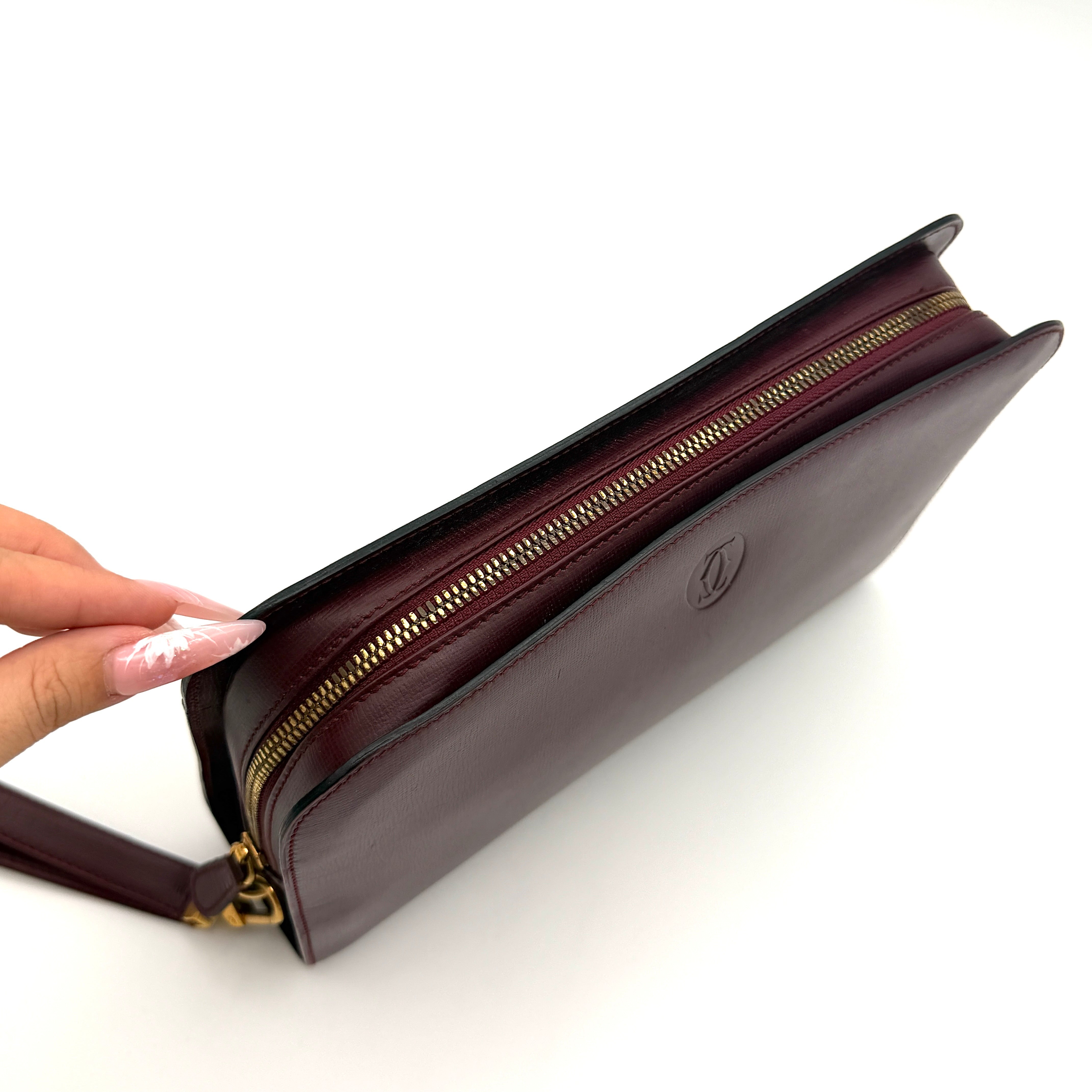 Cartier Must de Cartier clutch in burgundy leather with gold-tone zip and wrist strap, shown against a white background.