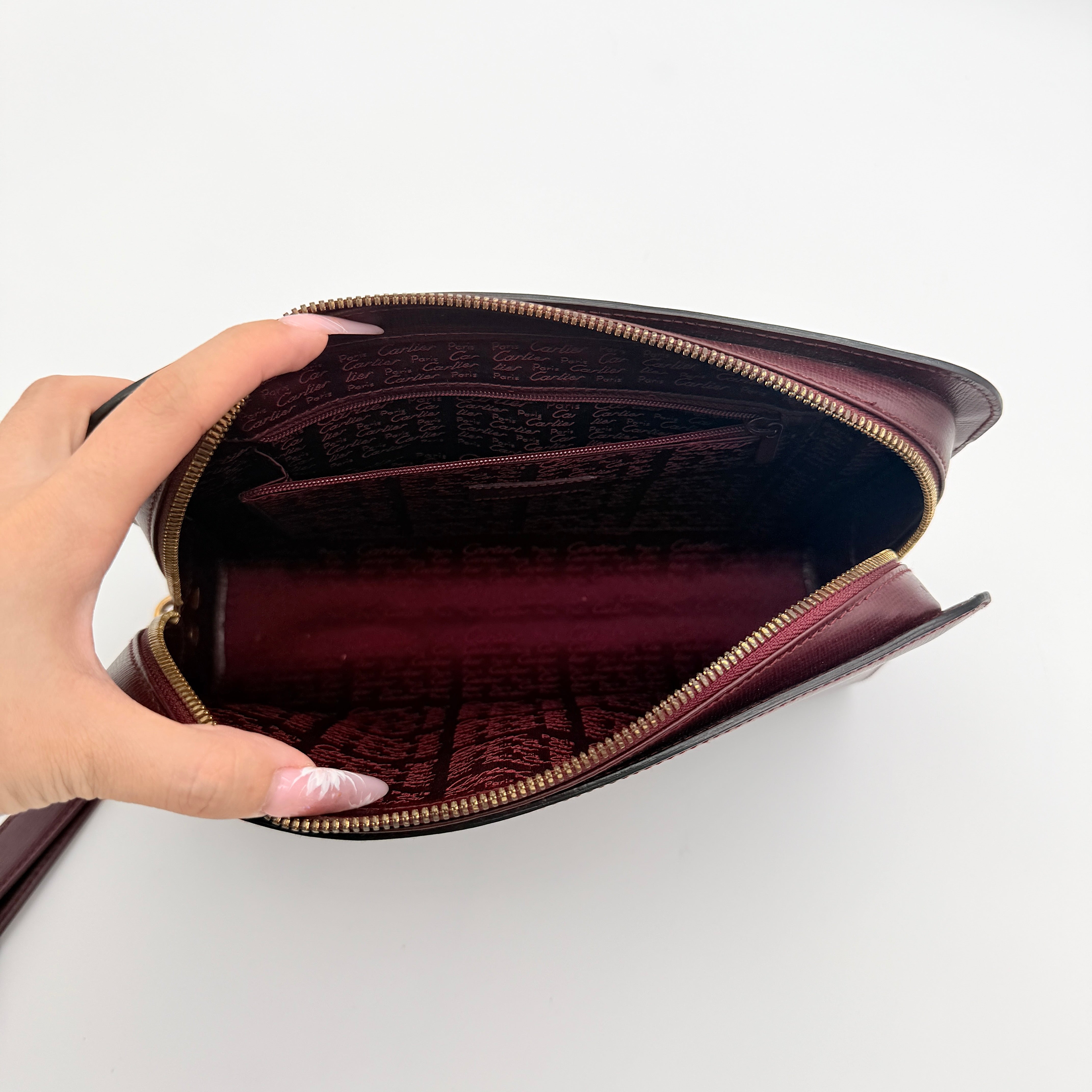 Cartier Must de Cartier clutch in burgundy leather with gold-tone zip and wrist strap, shown against a white background.