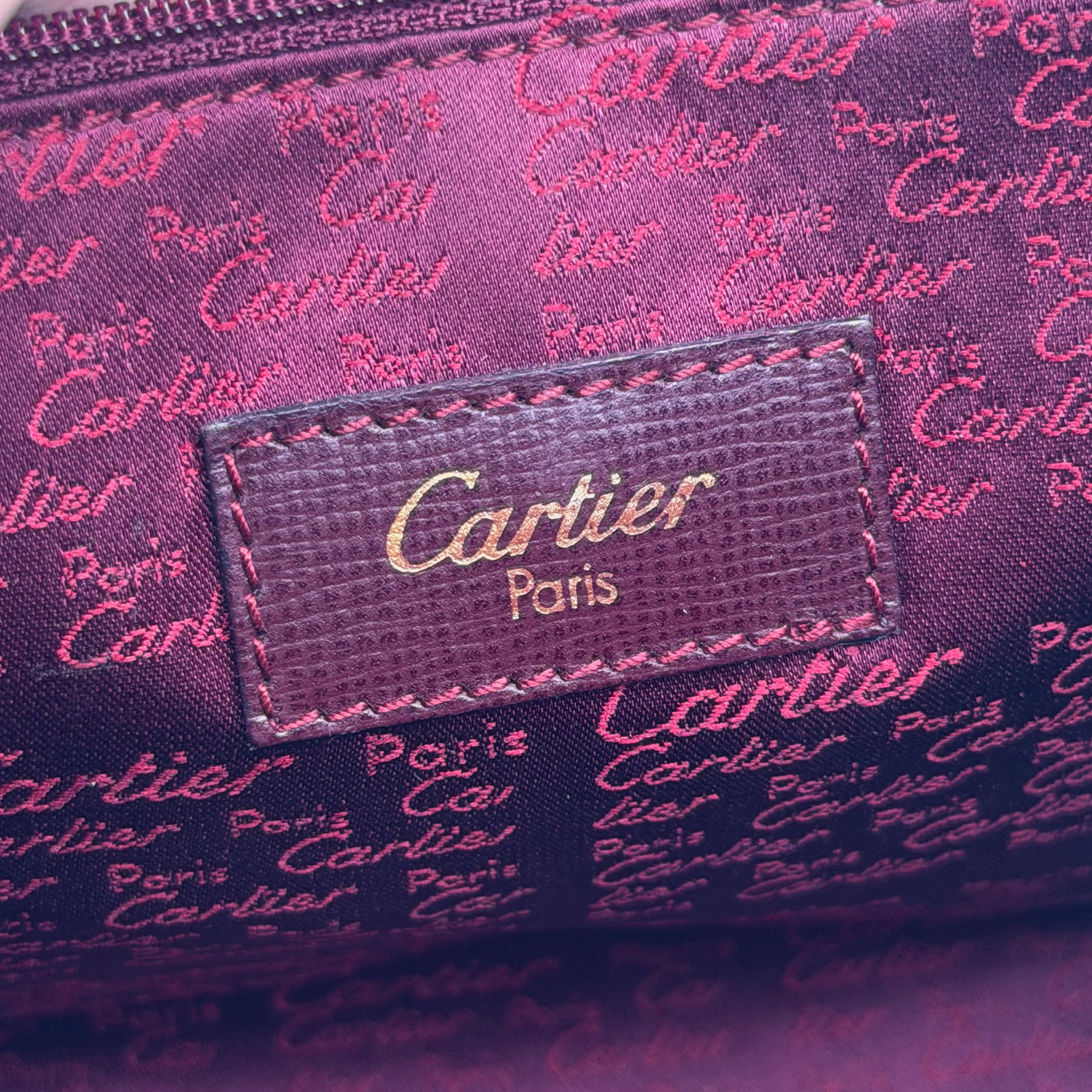 Cartier Must de Cartier clutch in burgundy leather with gold-tone zip and wrist strap, shown against a white background.