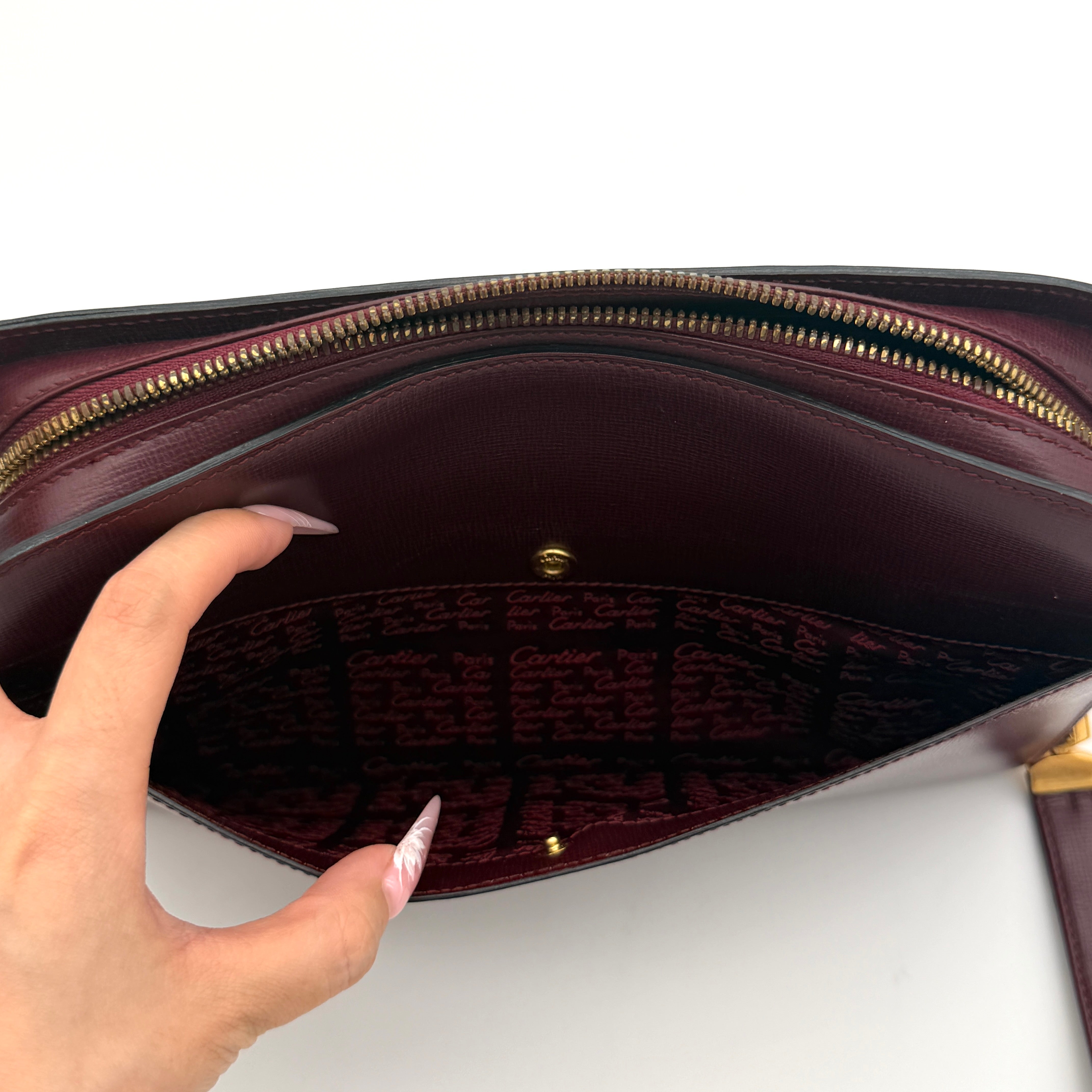Cartier Must de Cartier clutch in burgundy leather with gold-tone zip and wrist strap, shown against a white background.