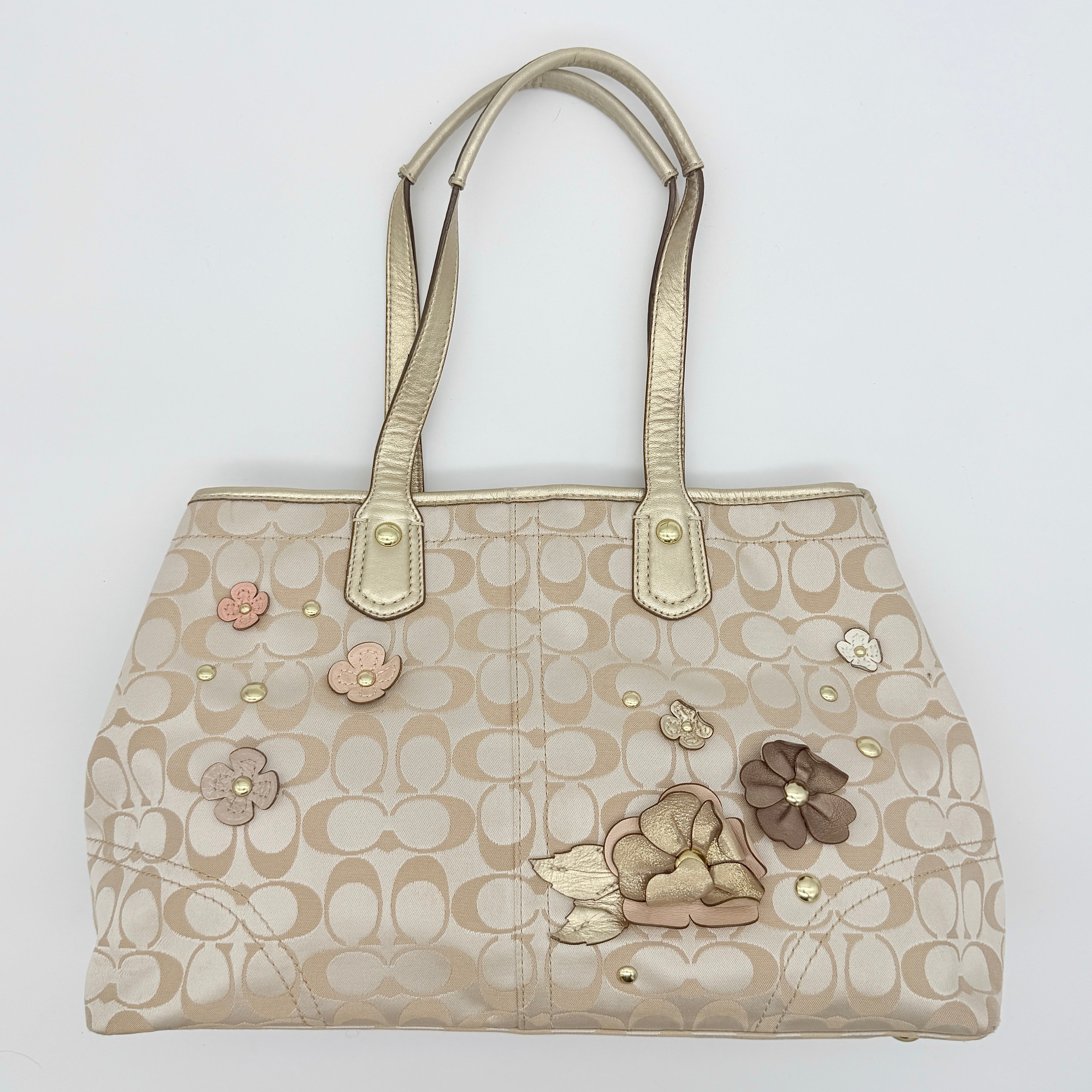 Coach beige carryall tote with floral appliqué and gold-tone handles, styled on a white background.