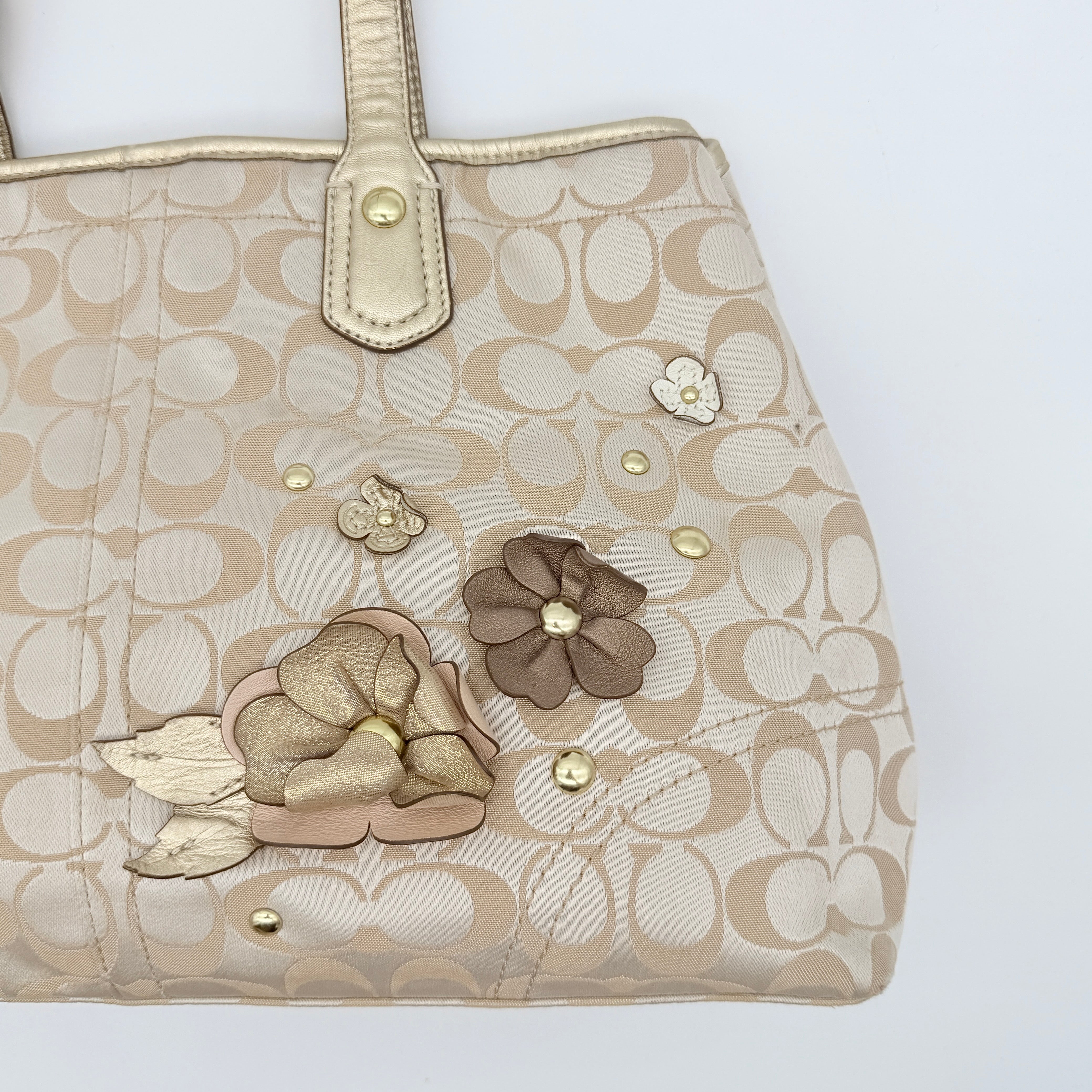 Coach beige carryall tote with floral appliqué and gold-tone handles, styled on a white background.