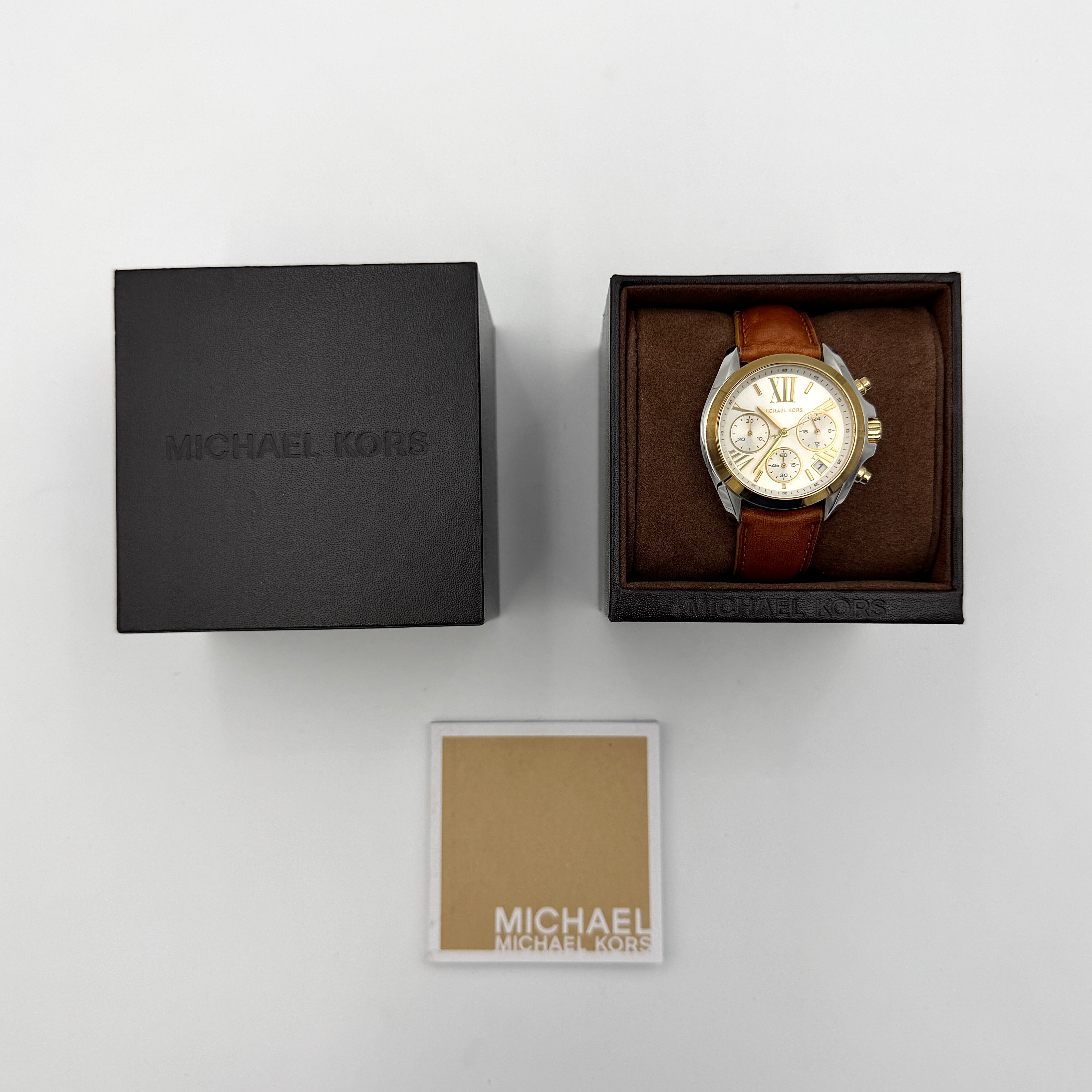 Michael Kors MK-2301 gold and silver tone chronograph watch with brown leather strap, showcased on a brown cushion atop the branded box.