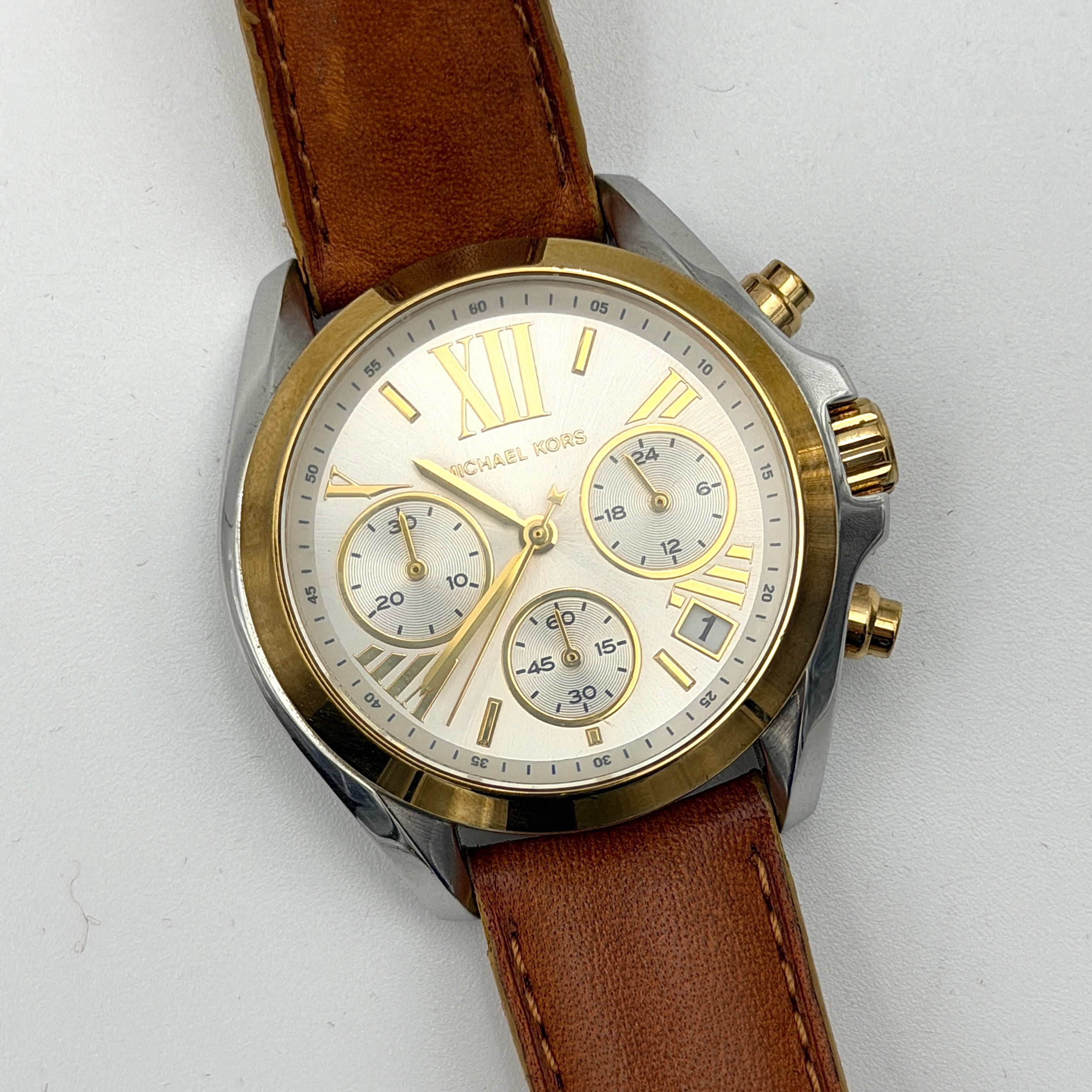 Michael Kors MK-2301 gold and silver tone chronograph watch with brown leather strap, showcased on a brown cushion atop the branded box.