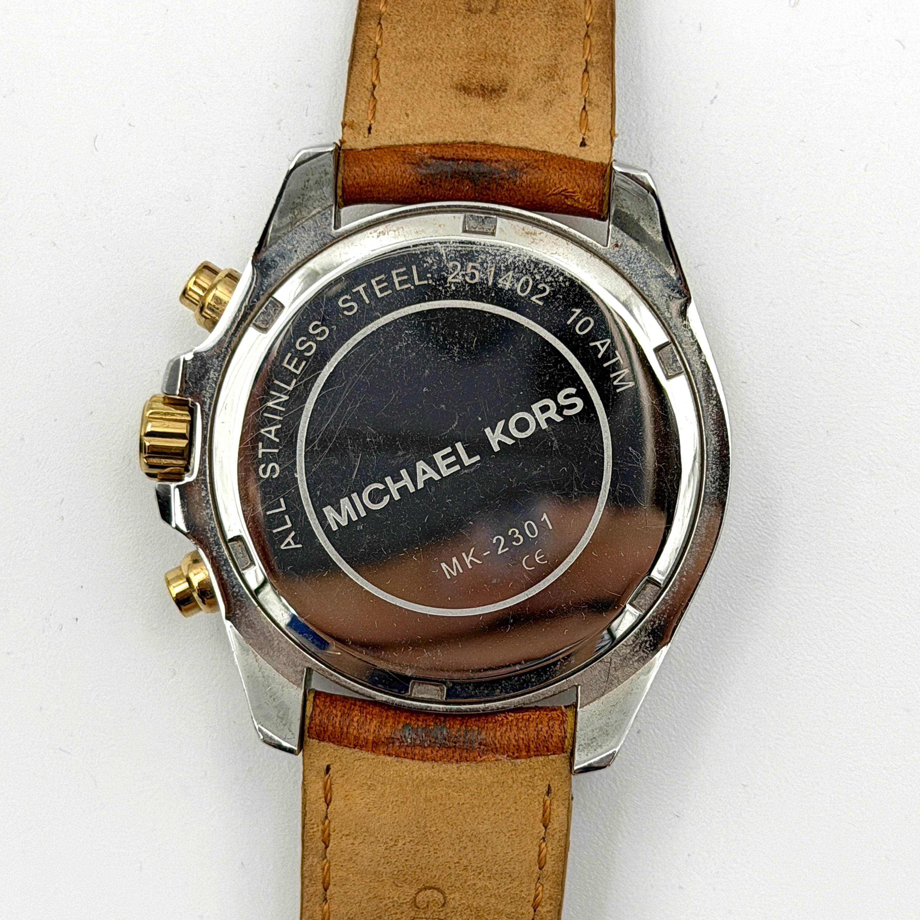 Michael Kors MK-2301 gold and silver tone chronograph watch with brown leather strap, showcased on a brown cushion atop the branded box.