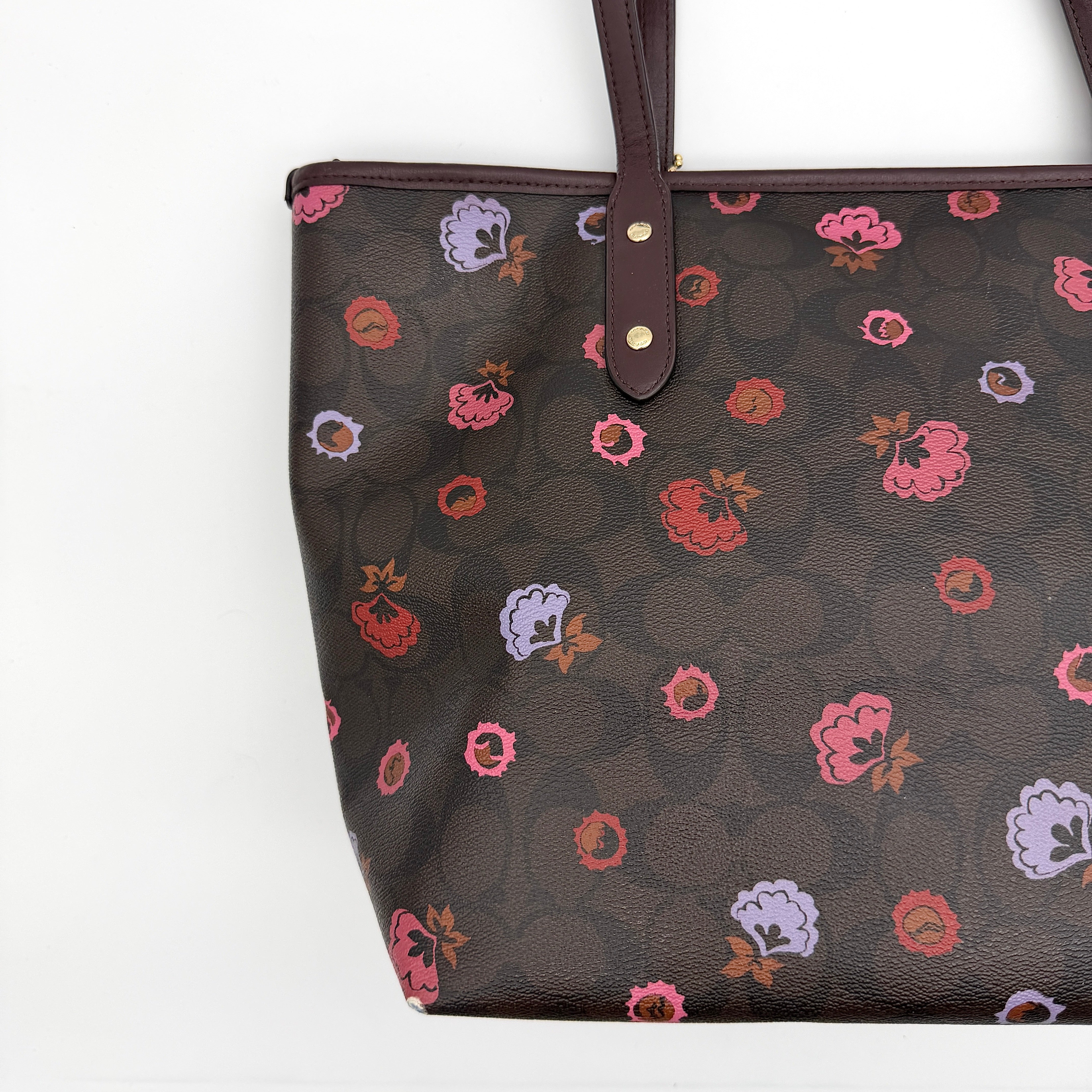 Coach brown coated canvas city tote with pink and lavender floral print, burgundy handles, and gold-tone logo at the front.