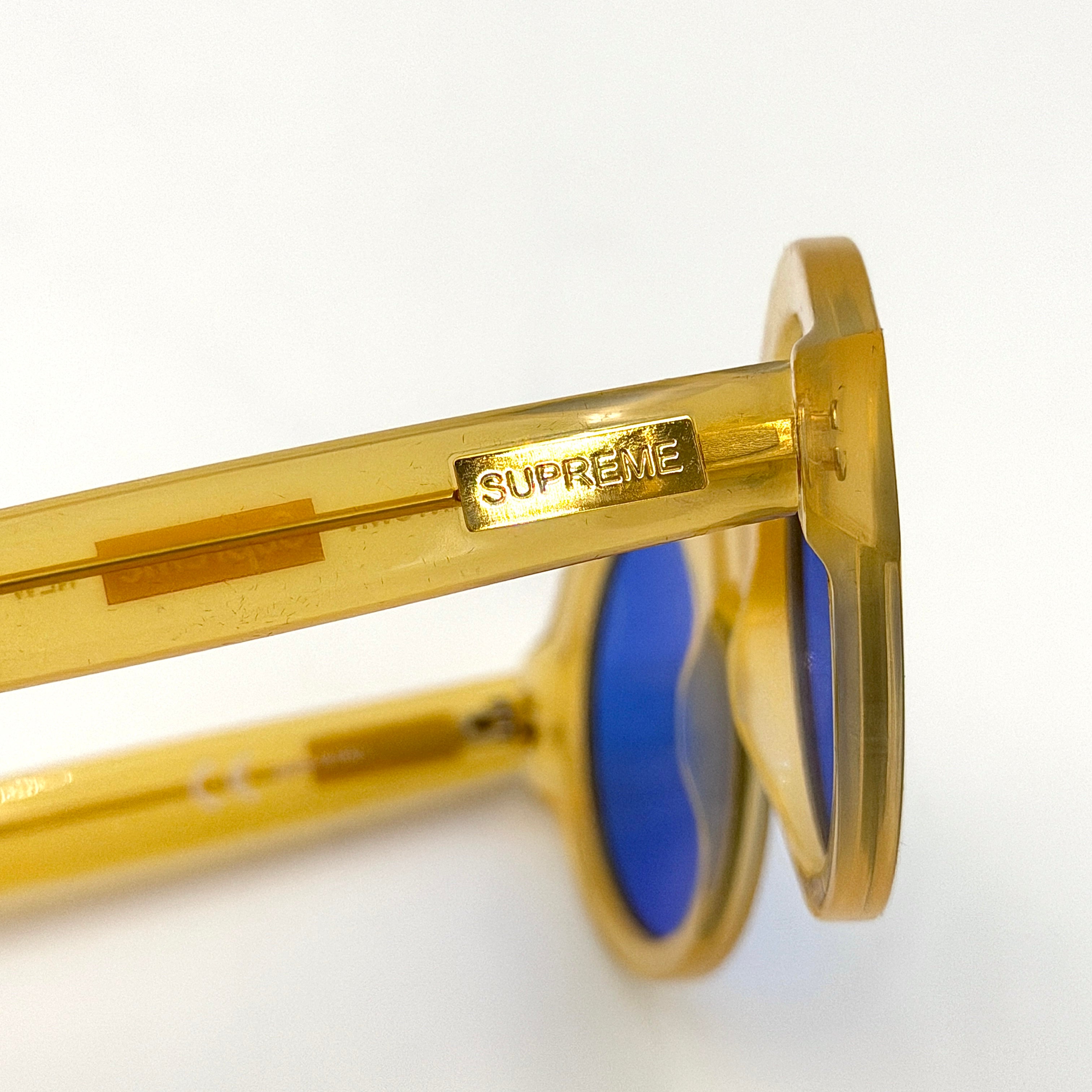 Supreme Downtown Sunglasses SS21 in translucent gold with vivid blue round lenses, set before a black Supreme case.