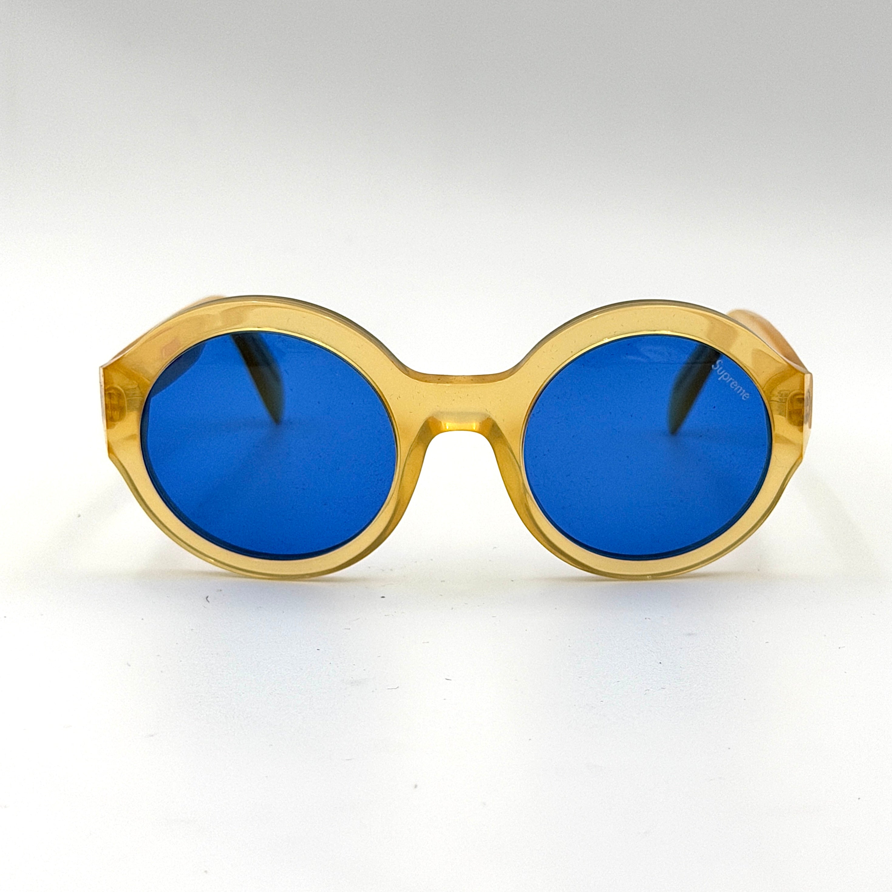 Supreme Downtown Sunglasses SS21 in translucent gold with vivid blue round lenses, set before a black Supreme case.