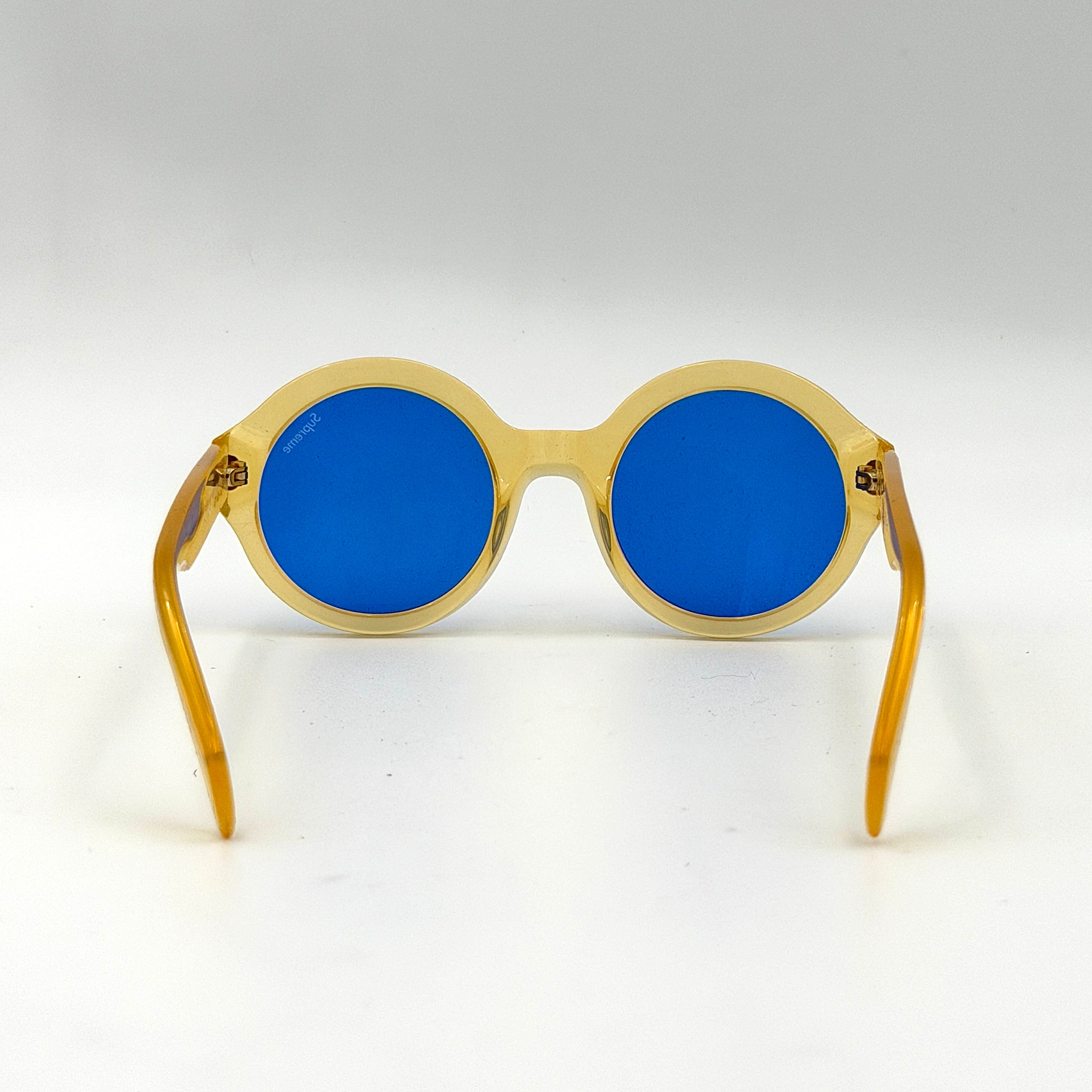 Supreme Downtown Sunglasses SS21 in translucent gold with vivid blue round lenses, set before a black Supreme case.