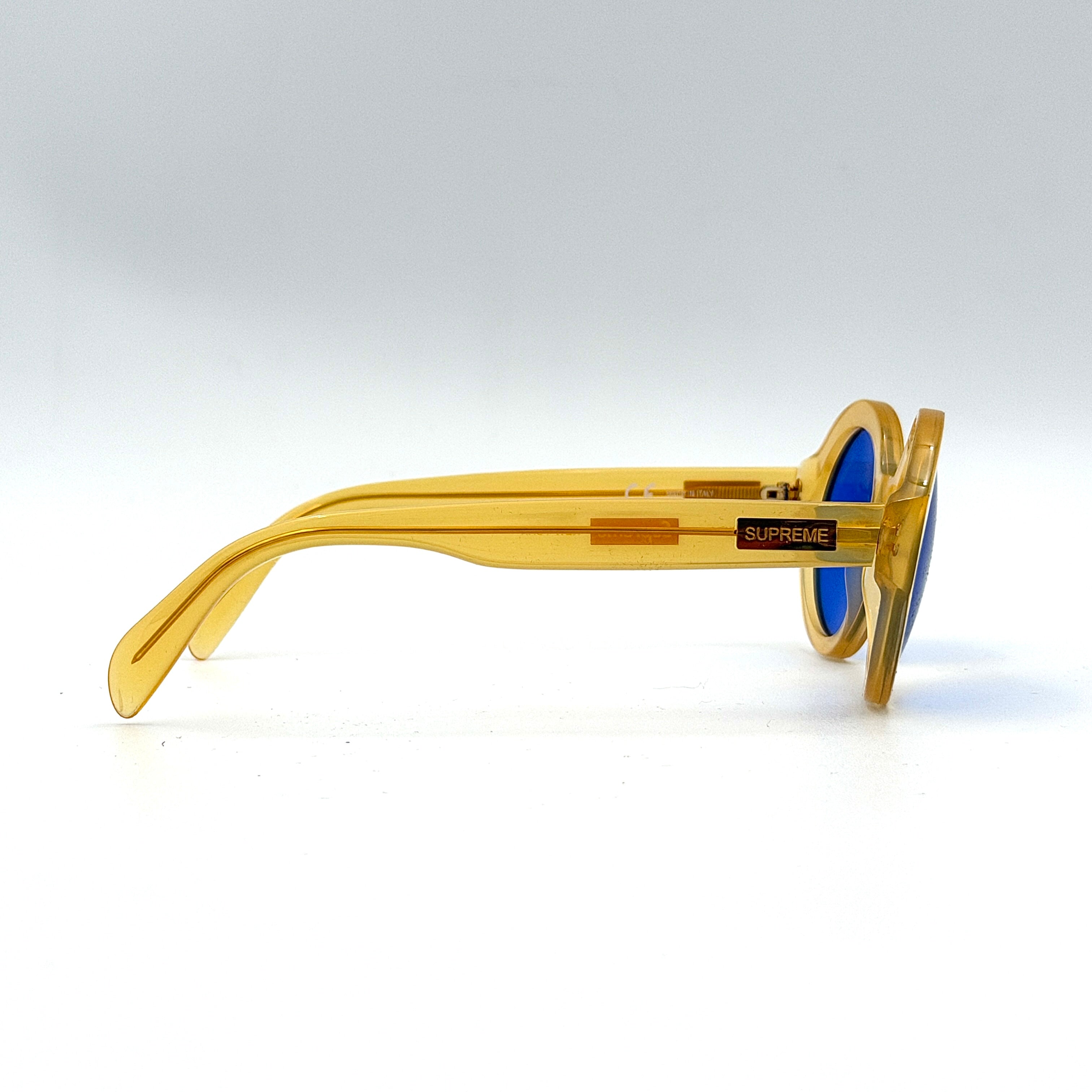 Supreme Downtown Sunglasses SS21 in translucent gold with vivid blue round lenses, set before a black Supreme case.