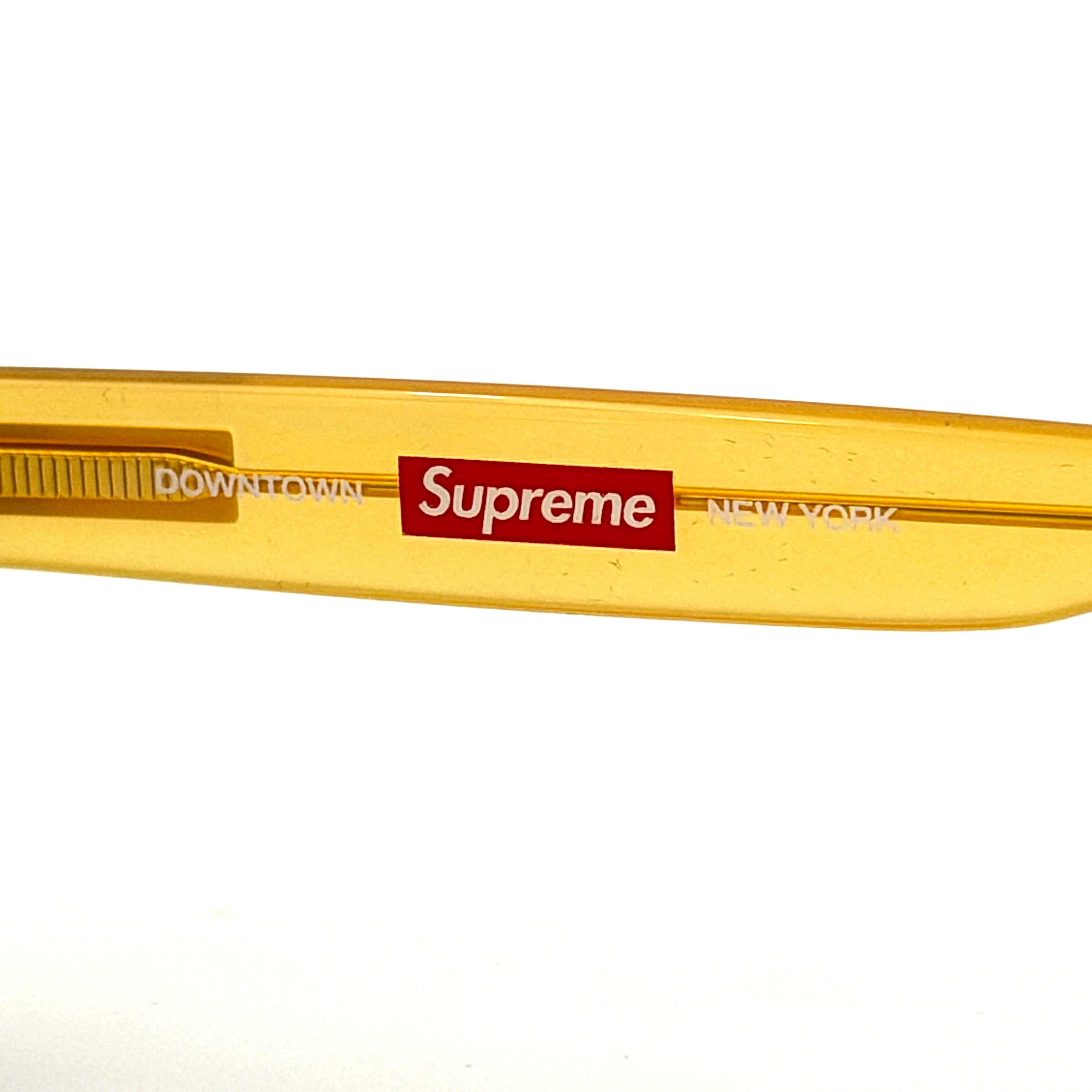 Supreme Downtown Sunglasses SS21 in translucent gold with vivid blue round lenses, set before a black Supreme case.