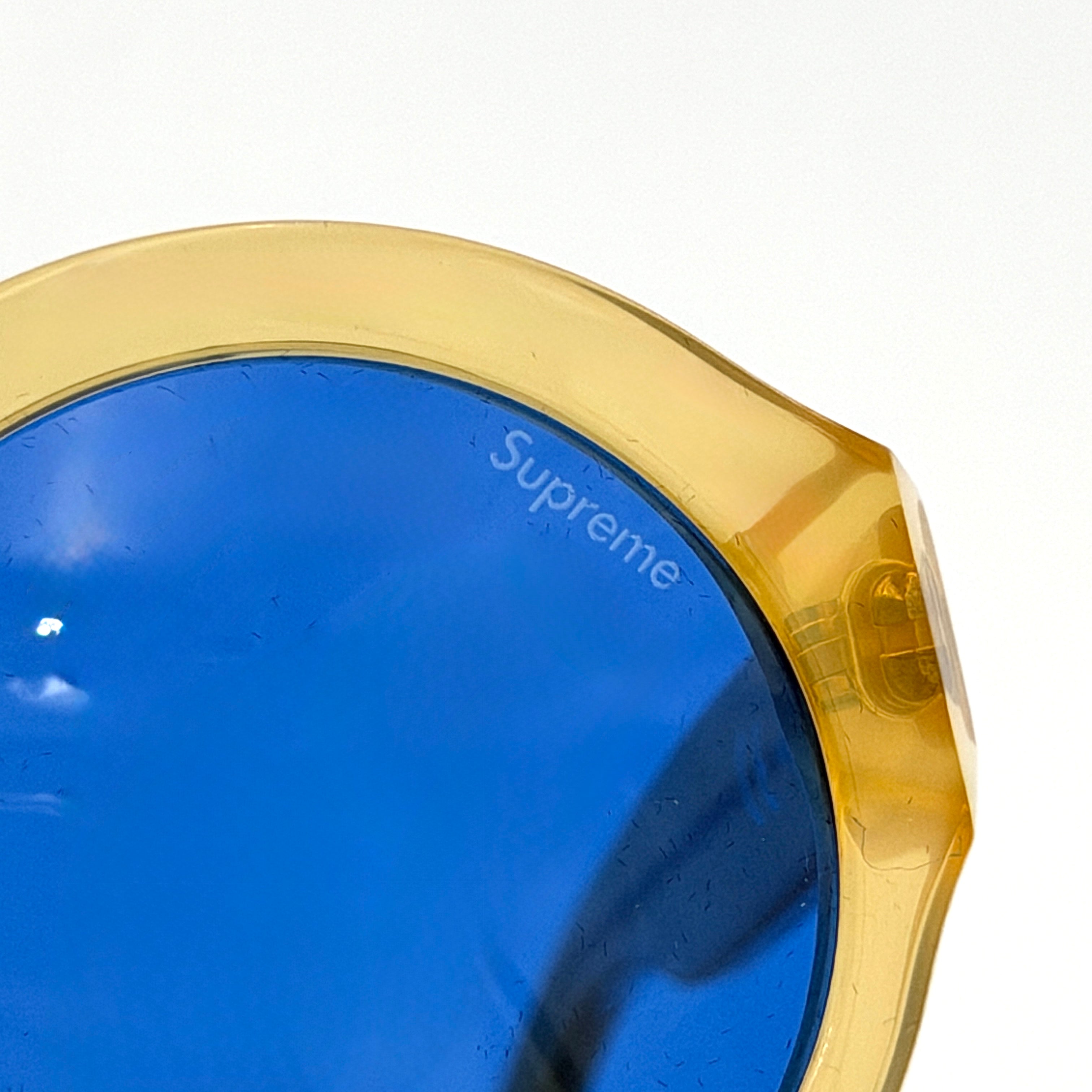Supreme Downtown Sunglasses SS21 in translucent gold with vivid blue round lenses, set before a black Supreme case.