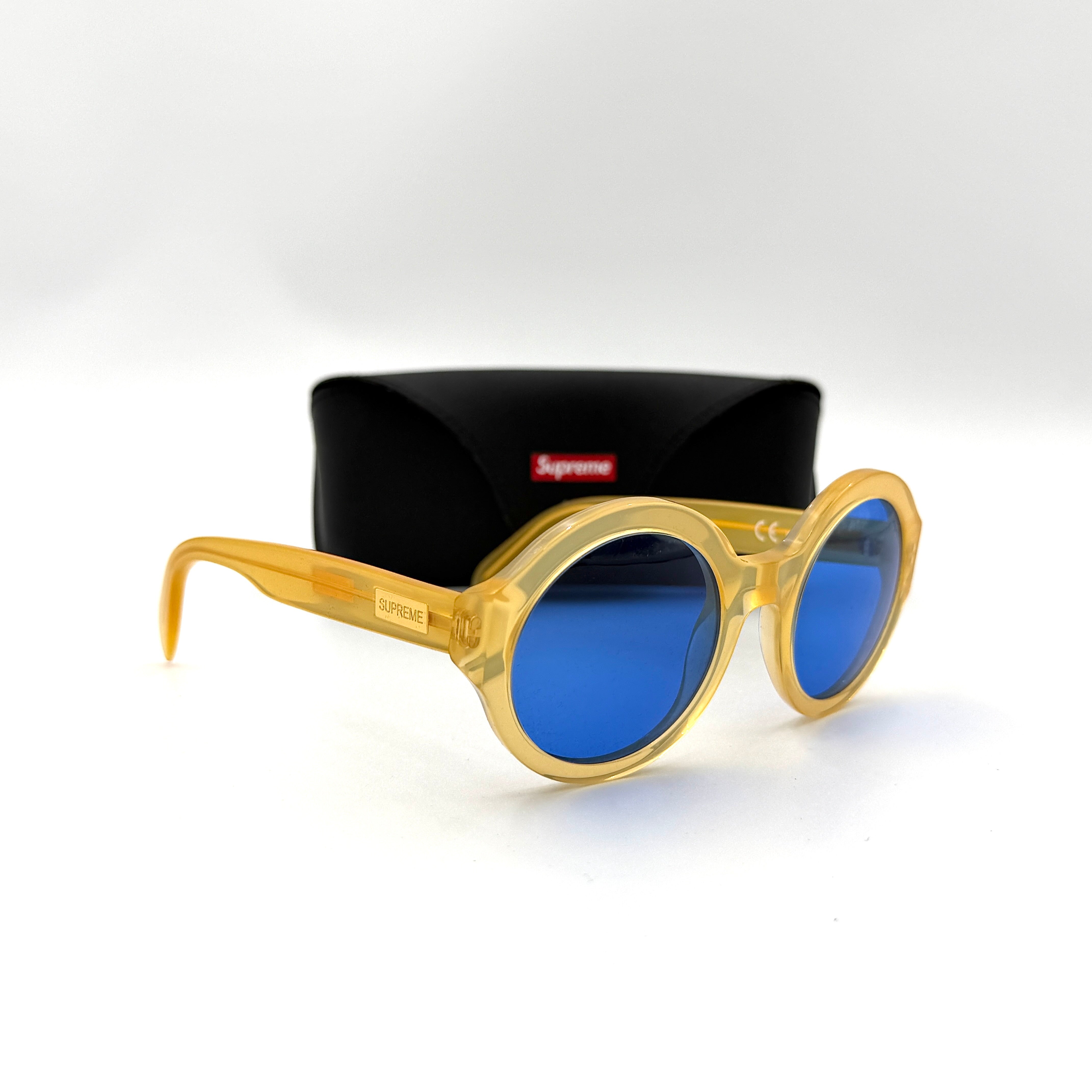 Supreme Downtown Sunglasses SS21 in translucent gold with vivid blue round lenses, set before a black Supreme case.