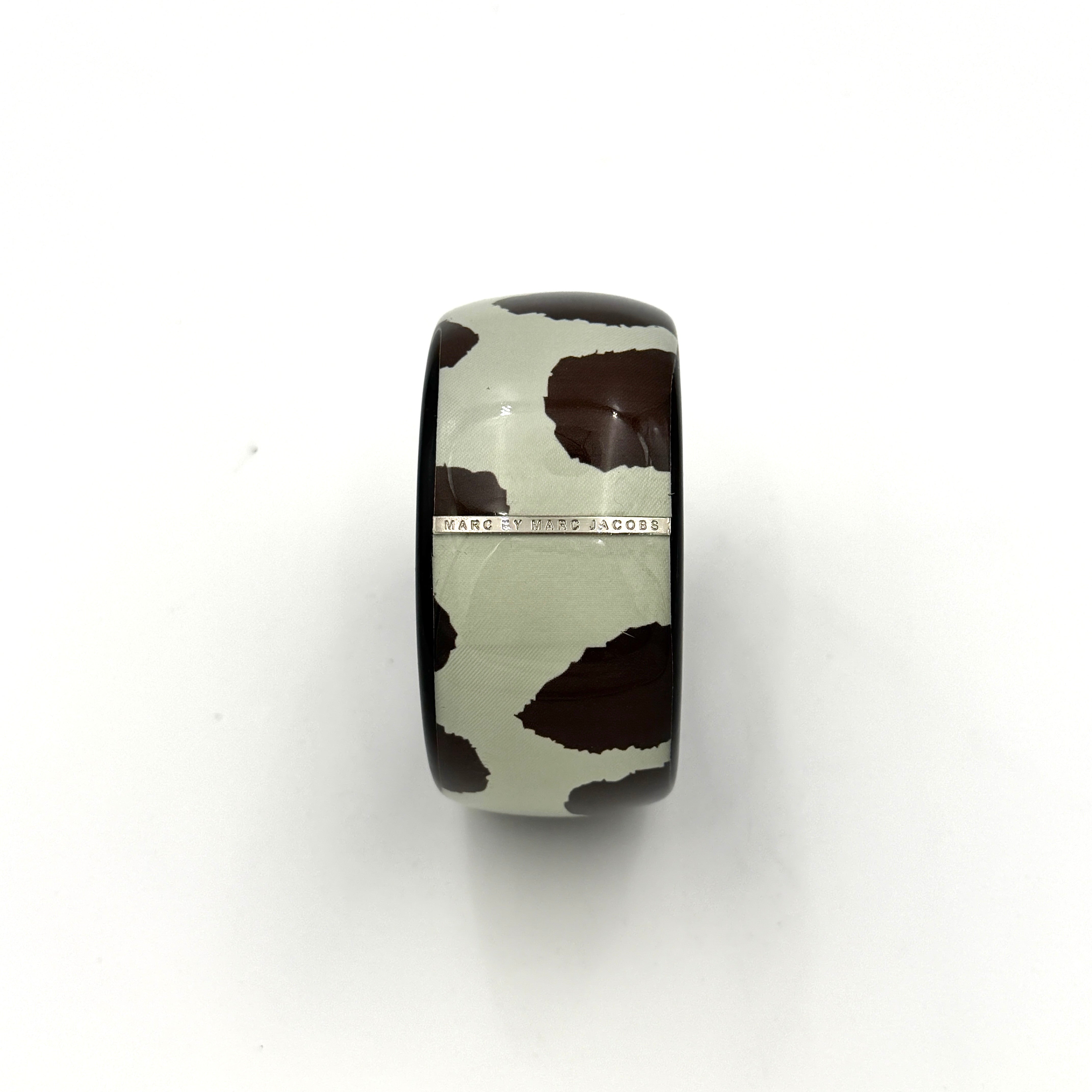 A wide Marc Jacobs bangle bracelet in sage green and dark brown cow print resin, featuring a slim silvertone logo plaque.