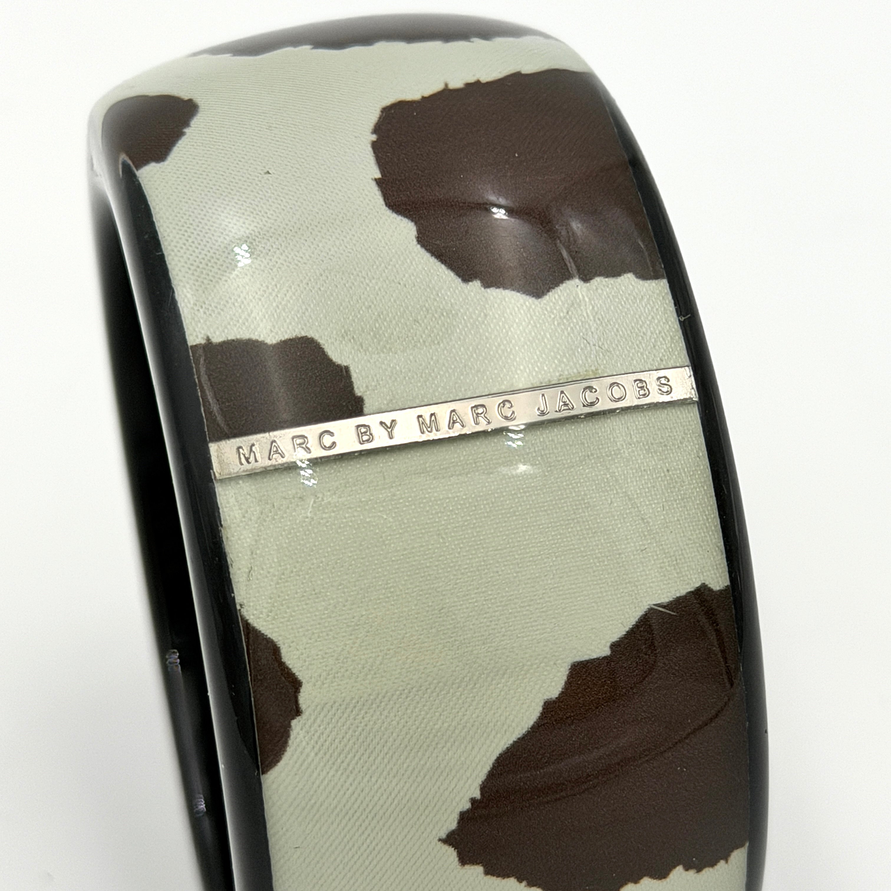 A wide Marc Jacobs bangle bracelet in sage green and dark brown cow print resin, featuring a slim silvertone logo plaque.