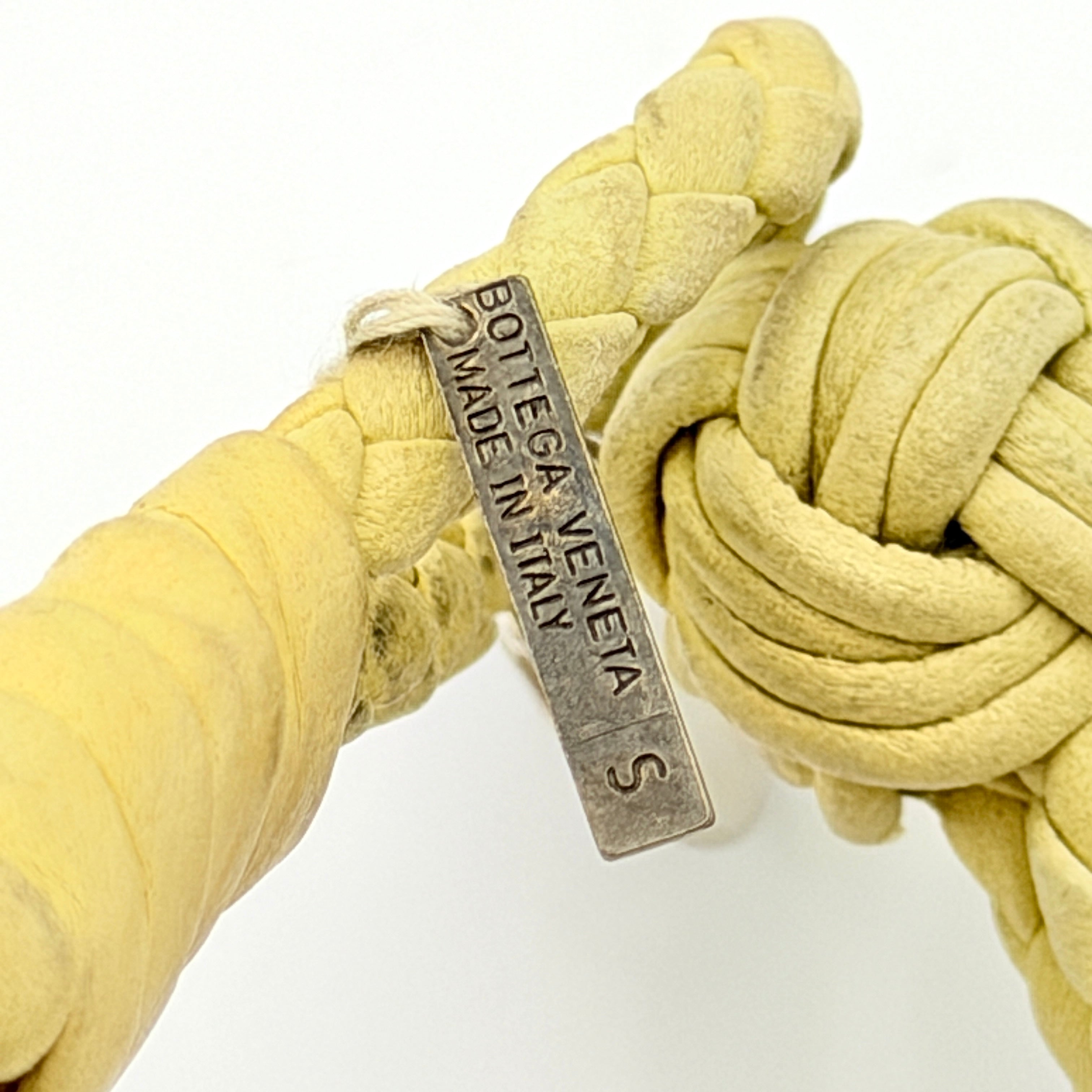 A braided pastel yellow Bottega Veneta Intrecciato leather bracelet with signature knot closure and engraved silver-tone tags.