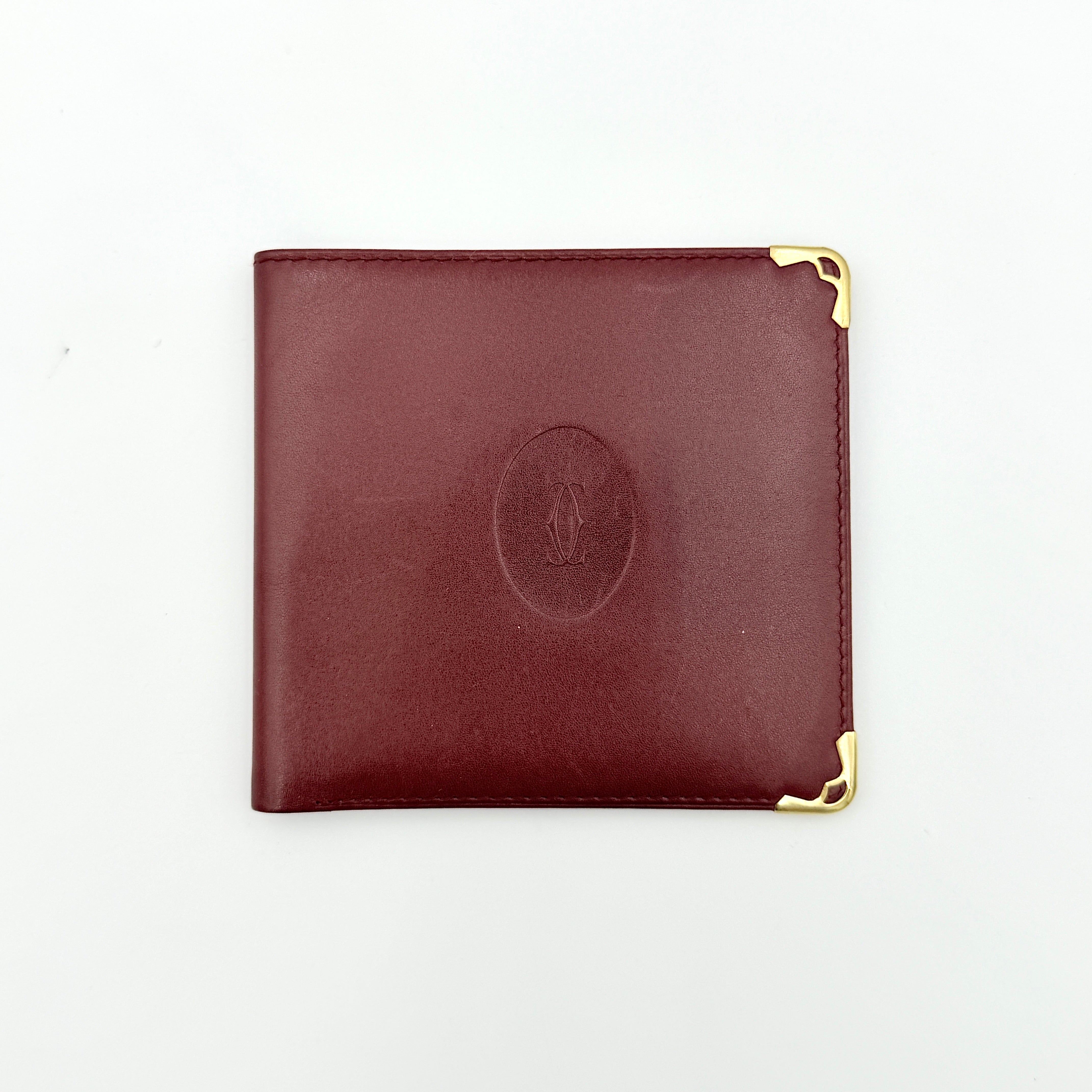 Cartier Must de Cartier leather wallet in rich burgundy with gold-tone corner accents and an embossed logo, photographed closed against a white background.