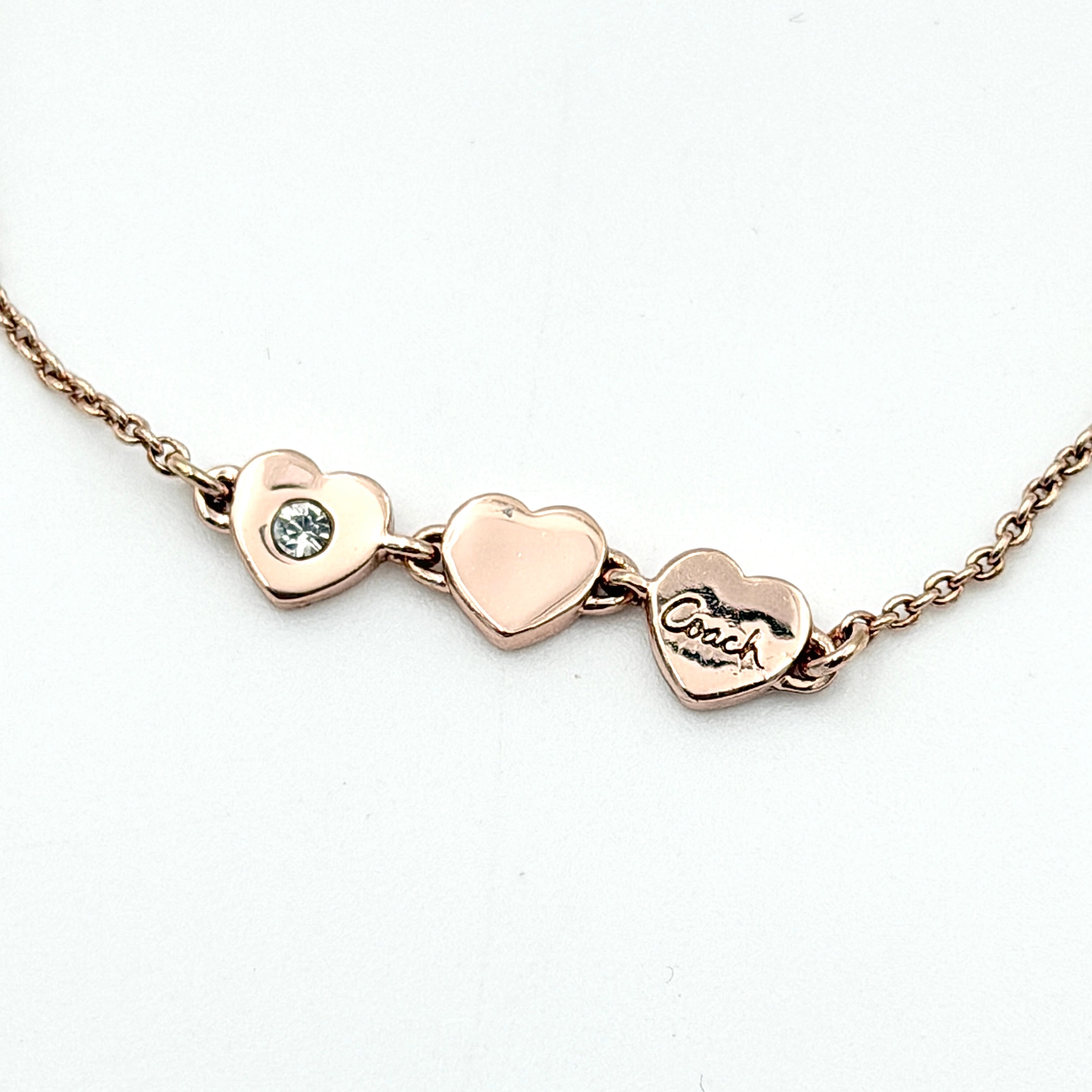 Coach gold bracelet featuring three heart-shaped charms, one with a crystal, one plain, and one engraved with the Coach logo.