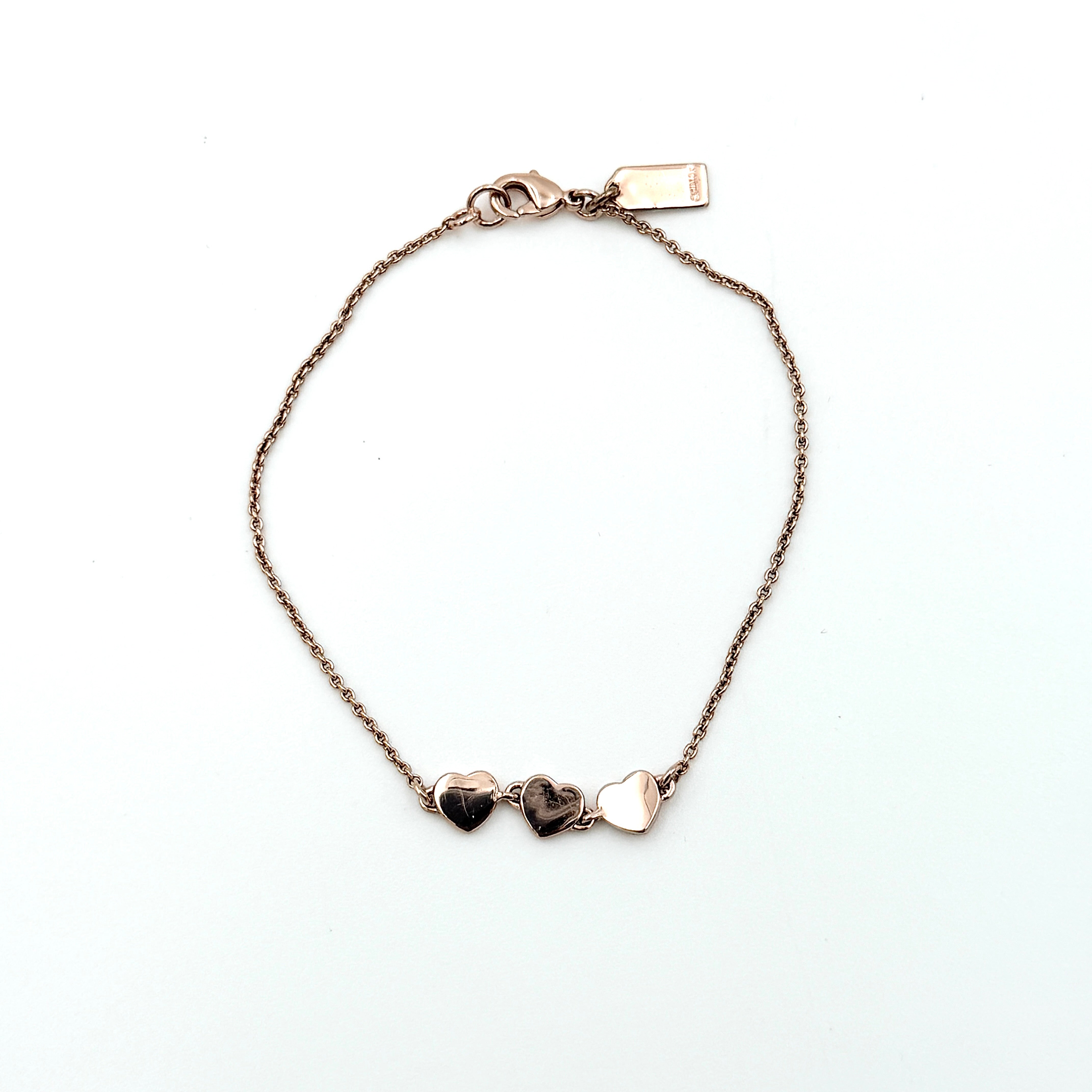 Coach gold bracelet featuring three heart-shaped charms, one with a crystal, one plain, and one engraved with the Coach logo.