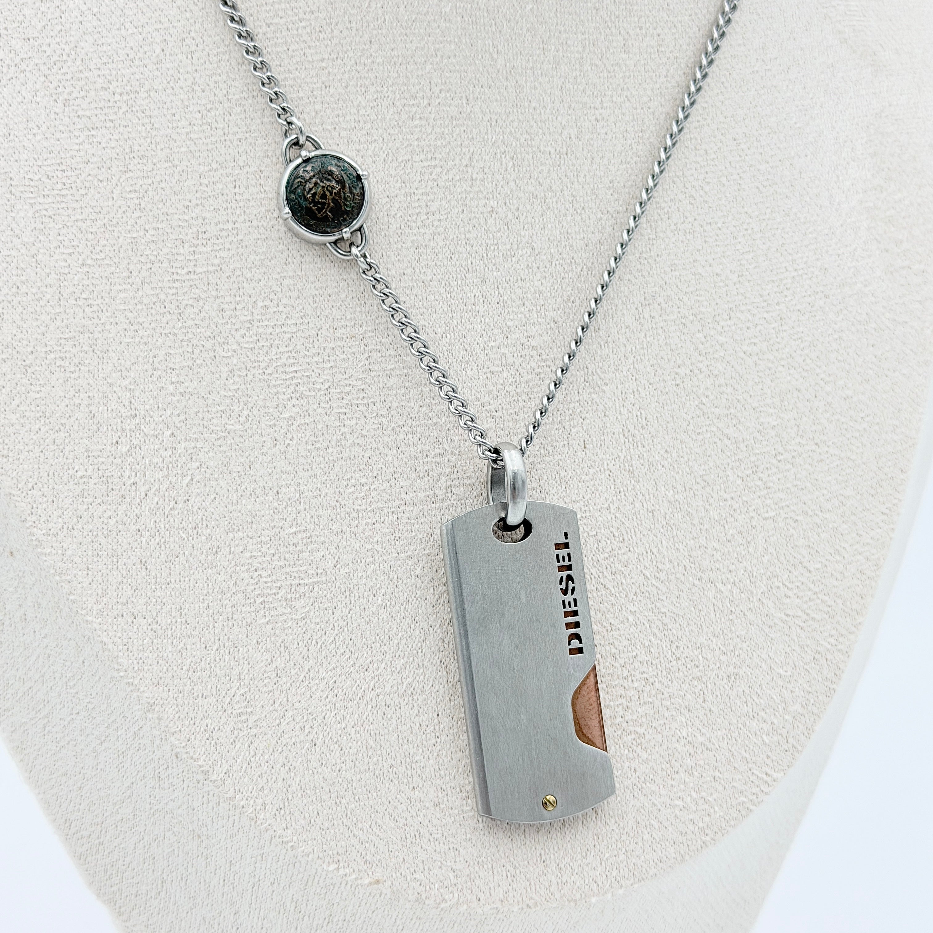 Diesel silver-tone stainless steel dog tag necklace with bronze accent, curb chain, and round detail showcased on a white background.