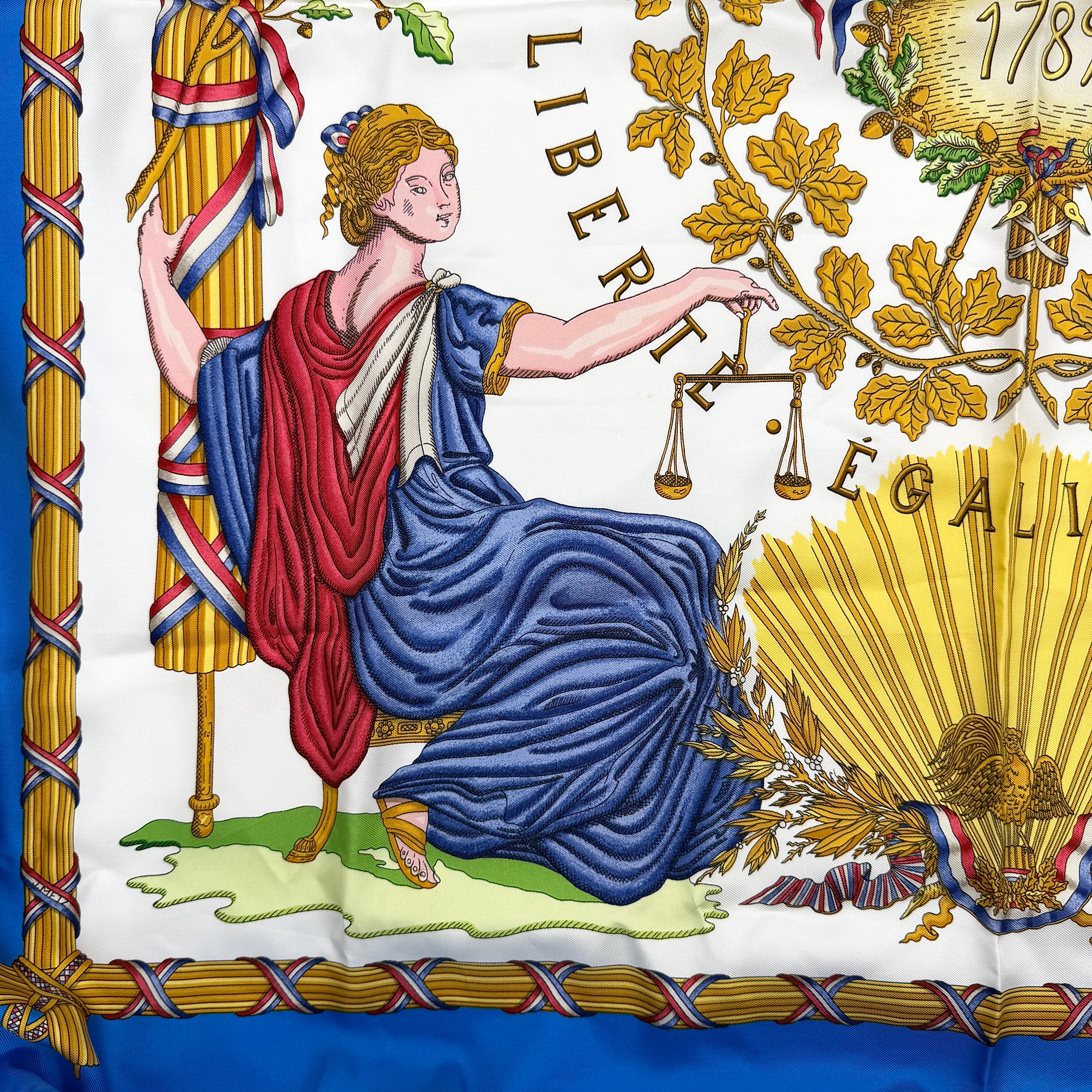 Hermès silk scarf with blue border, featuring a detailed French revolutionary motif and classical figures, shown with its signature orange box.
