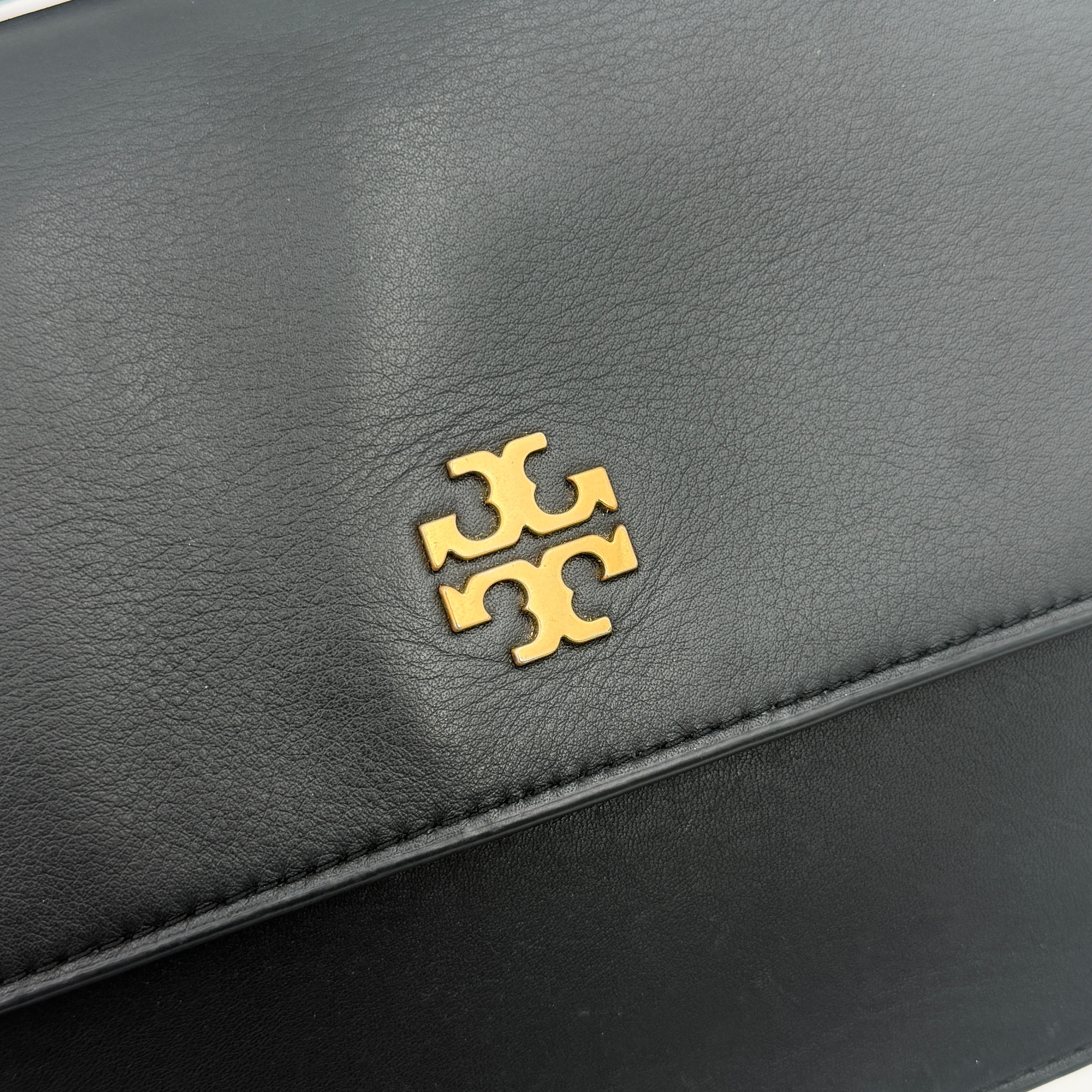 Tory Burch Brooke Crossbody Bag in black leather with gold-tone logo and a braided chain strap, elegantly displayed against a white backdrop.