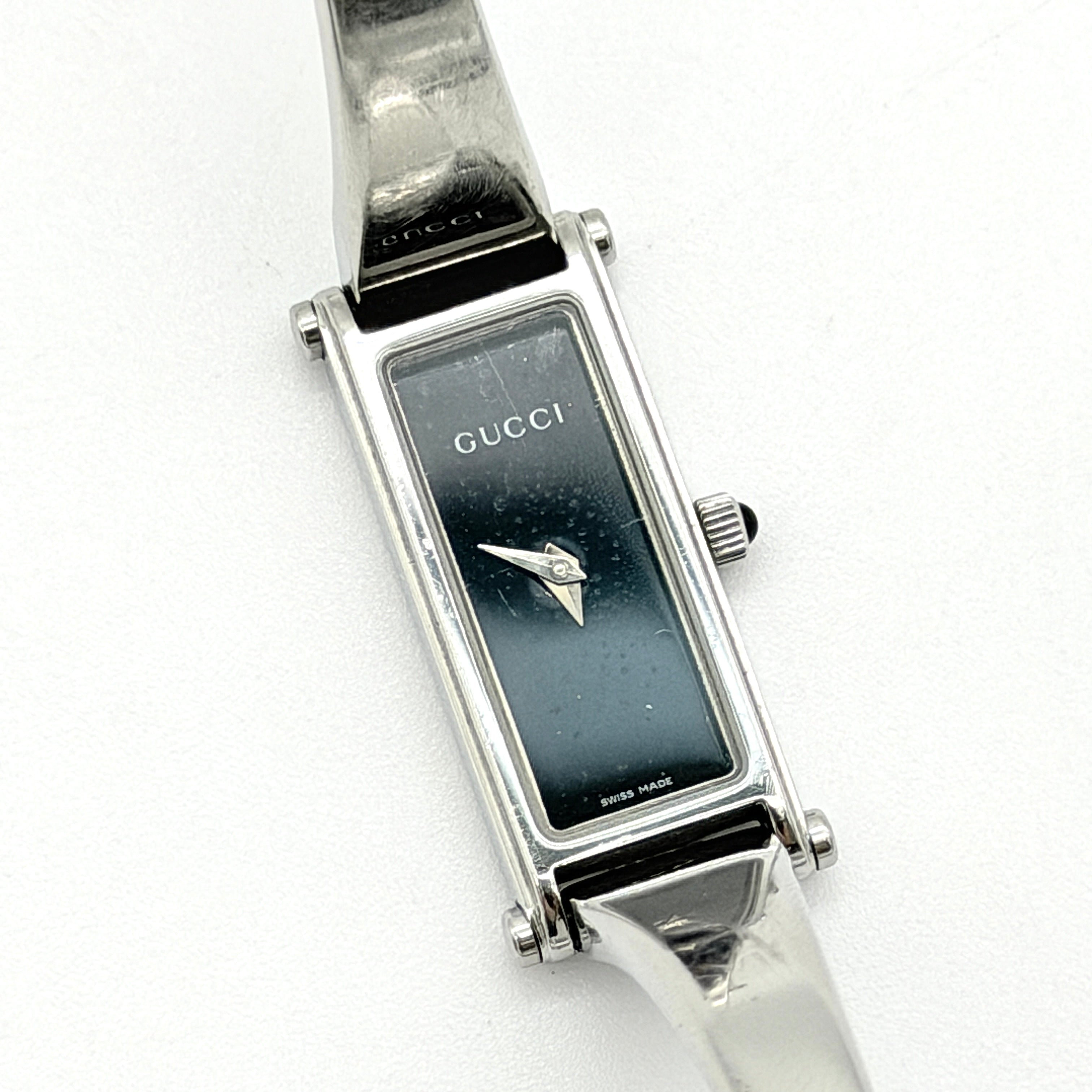 Gucci 1500L black dial bangle watch with stainless steel bracelet, photographed on a clear display stand.