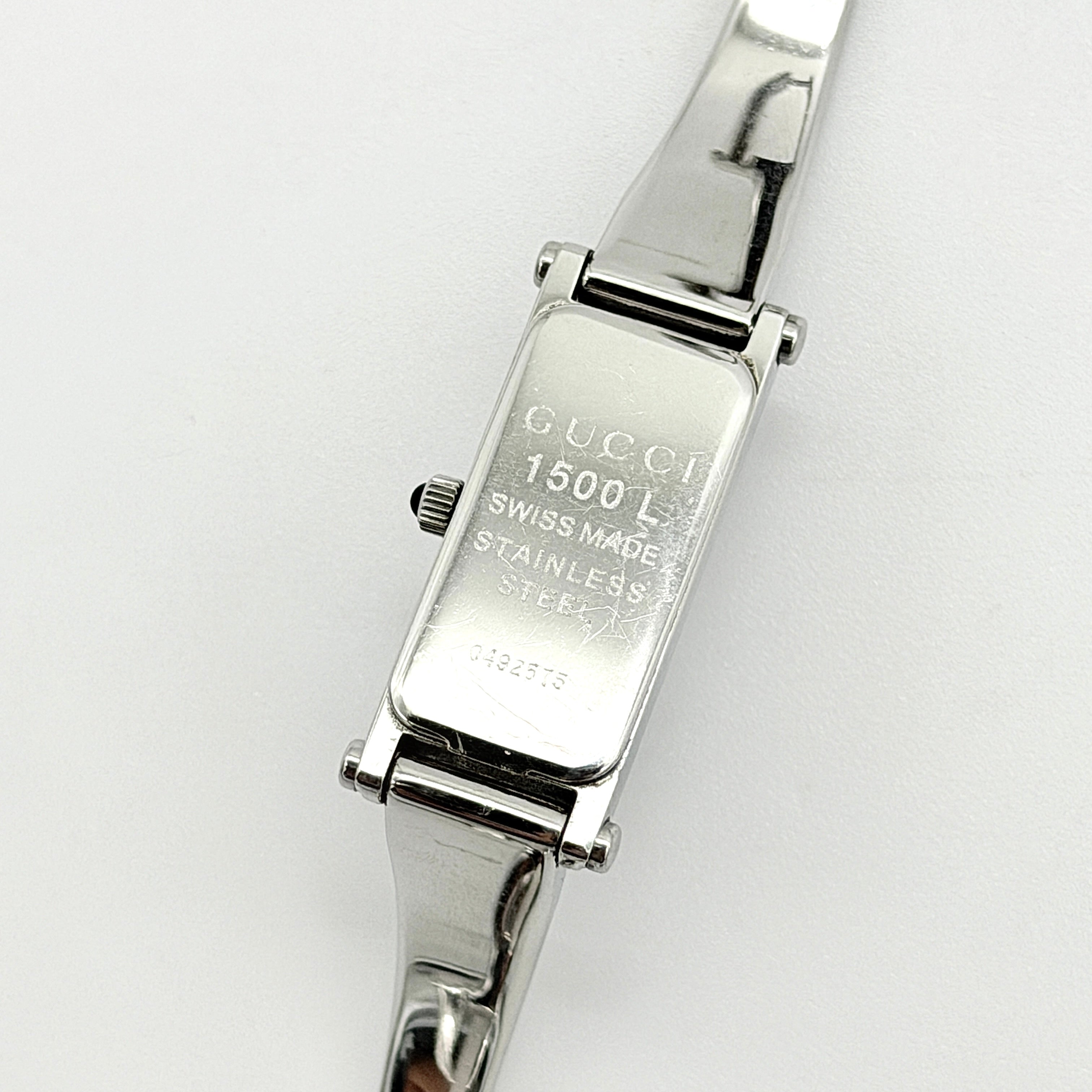 Gucci 1500L black dial bangle watch with stainless steel bracelet, photographed on a clear display stand.