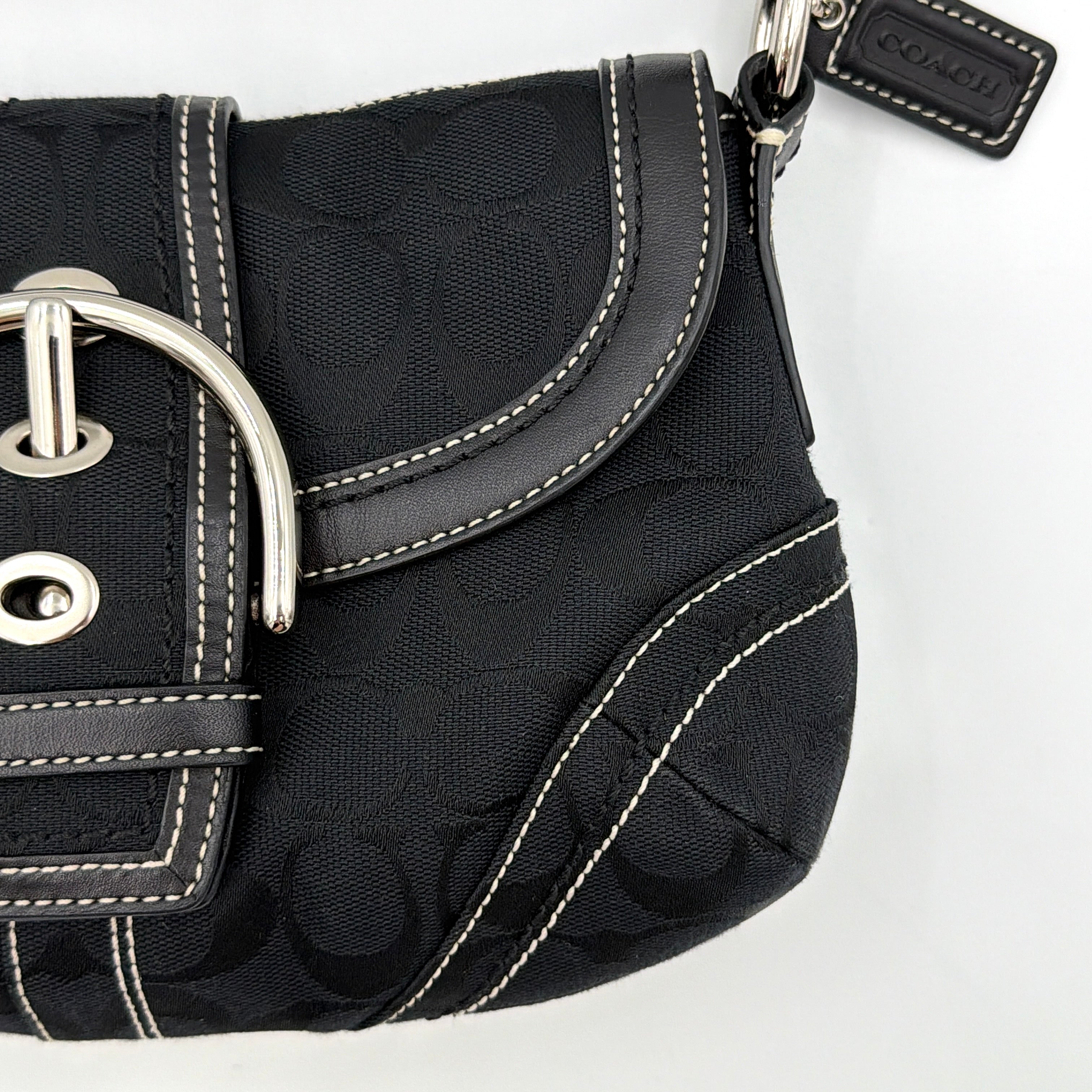 Coach black Signature Soho shoulder bag with oversized silver buckle, cream contrast stitching, and monogram jacquard canvas backdrop.