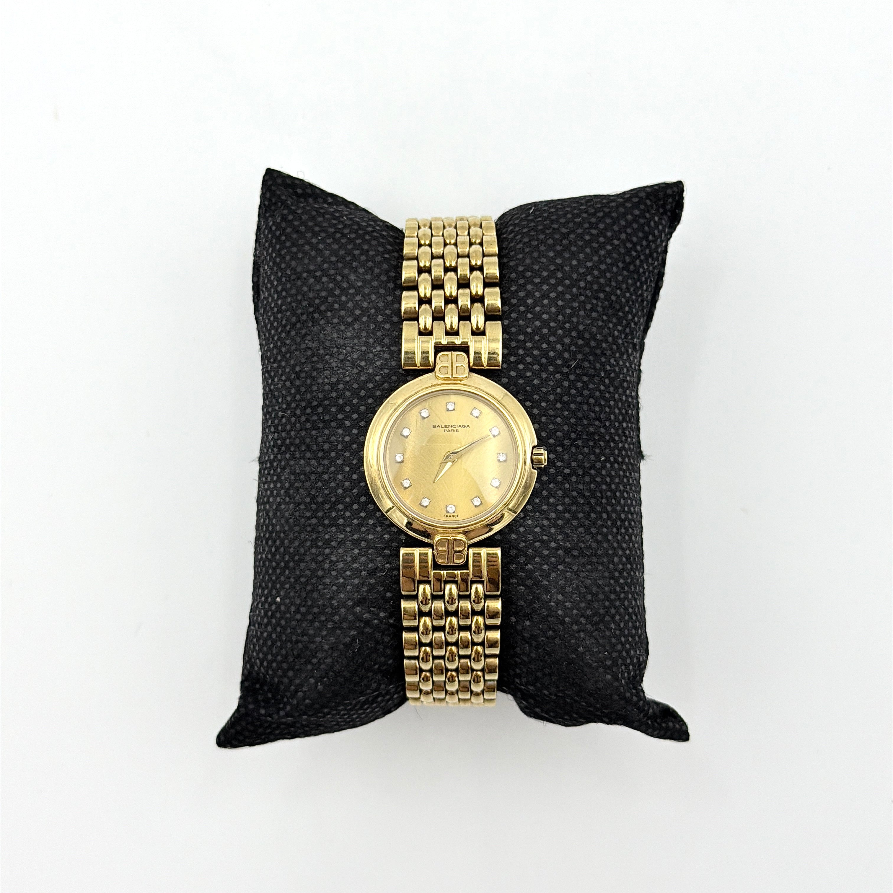 Balenciaga BB Quartz gold watch with a round dial, diamond-shaped hour markers, and a gold link bracelet displayed on a black cushion.