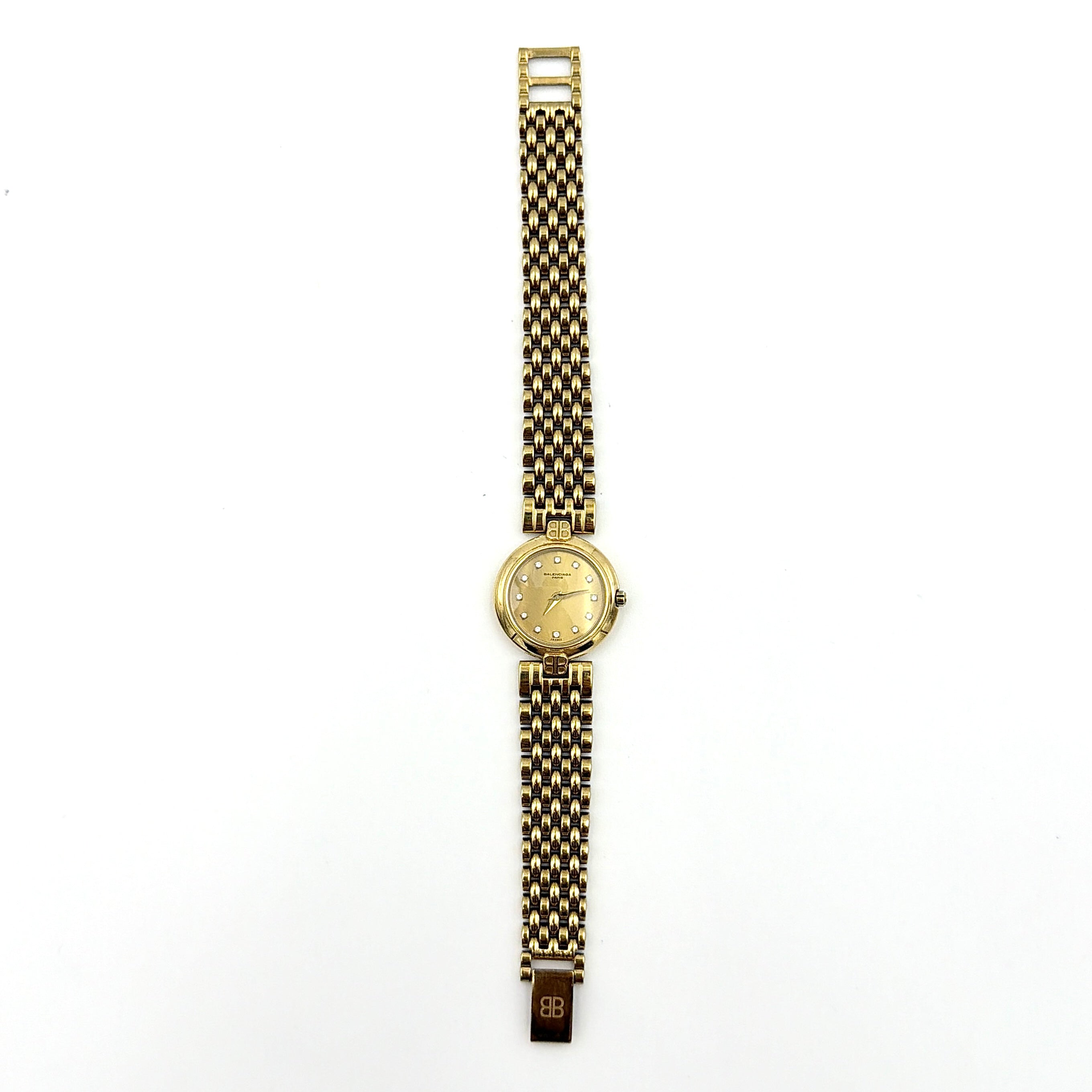 Balenciaga BB Quartz gold watch with a round dial, diamond-shaped hour markers, and a gold link bracelet displayed on a black cushion.