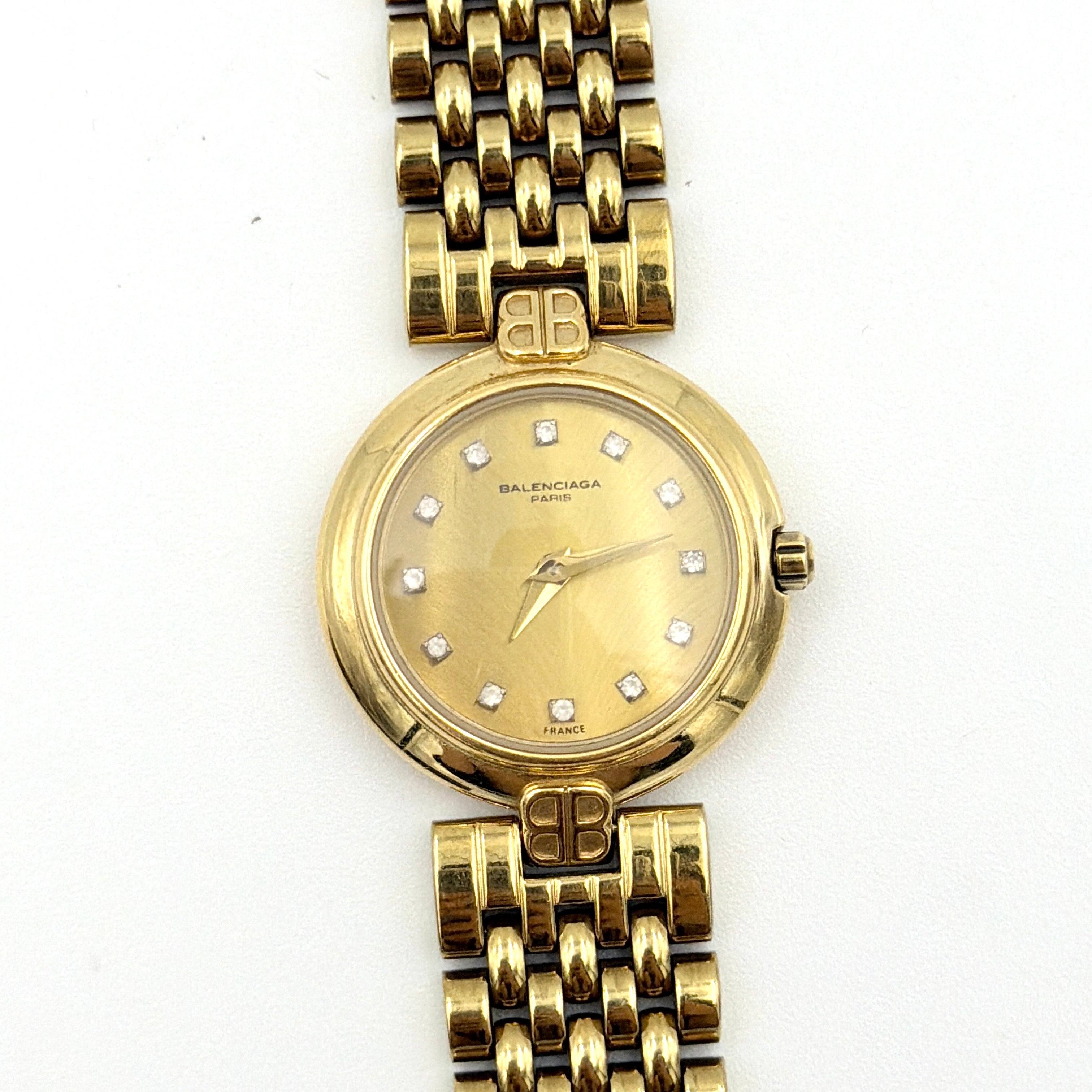 Balenciaga BB Quartz gold watch with a round dial, diamond-shaped hour markers, and a gold link bracelet displayed on a black cushion.