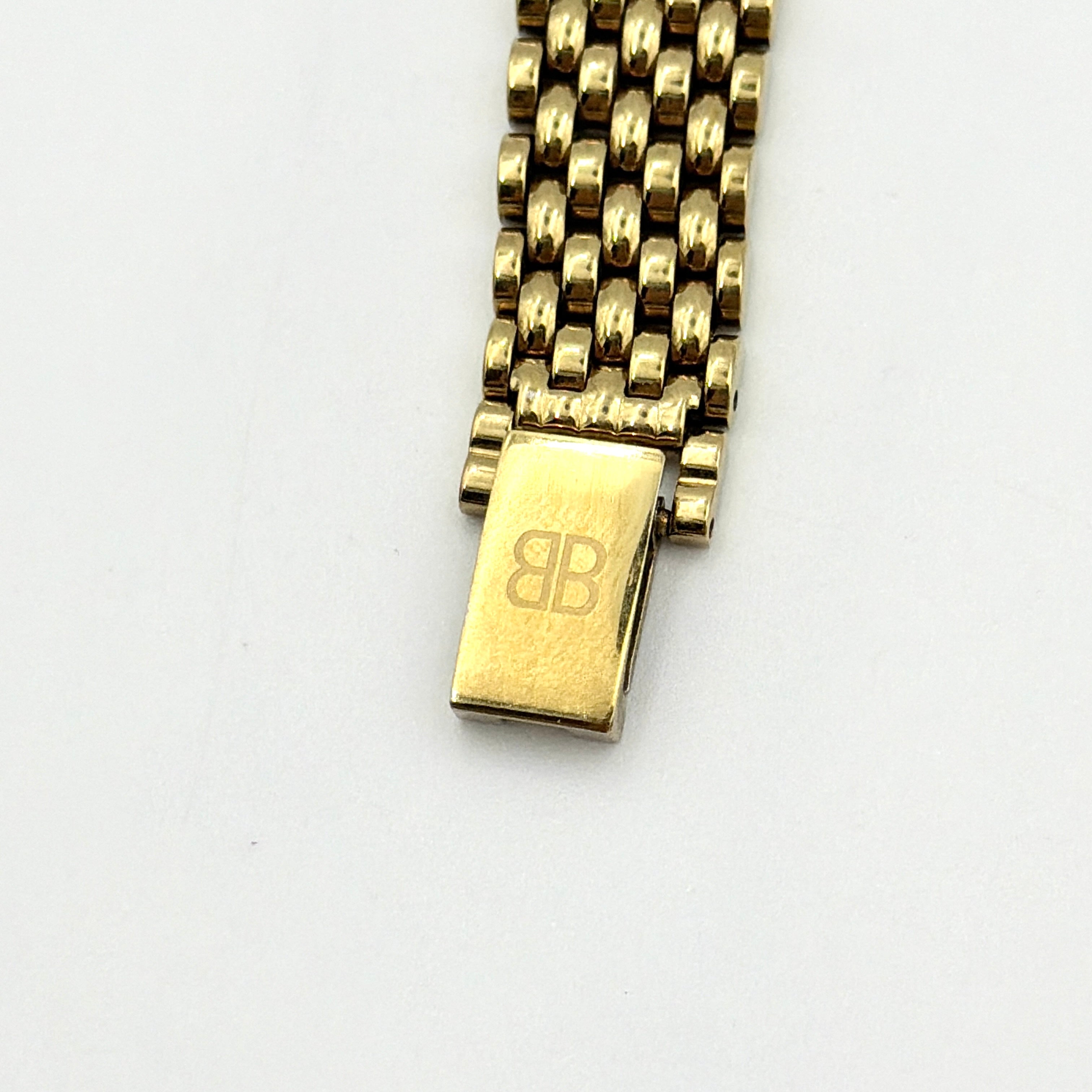 Balenciaga BB Quartz gold watch with a round dial, diamond-shaped hour markers, and a gold link bracelet displayed on a black cushion.