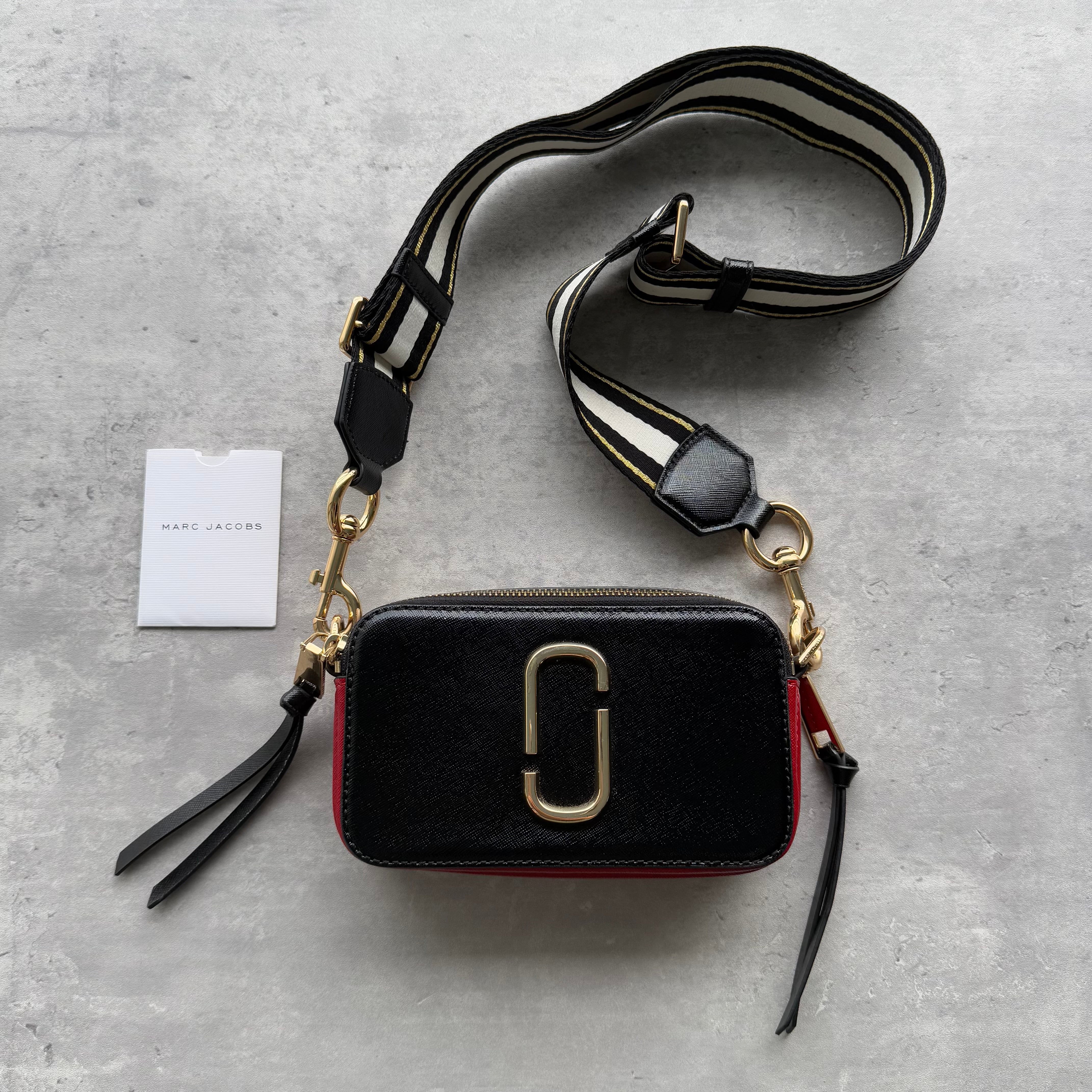 Marc Jacobs The Snapshot Crossbody Bag in black leather with gold double-J hardware, red side panel, and detachable striped webbing strap.