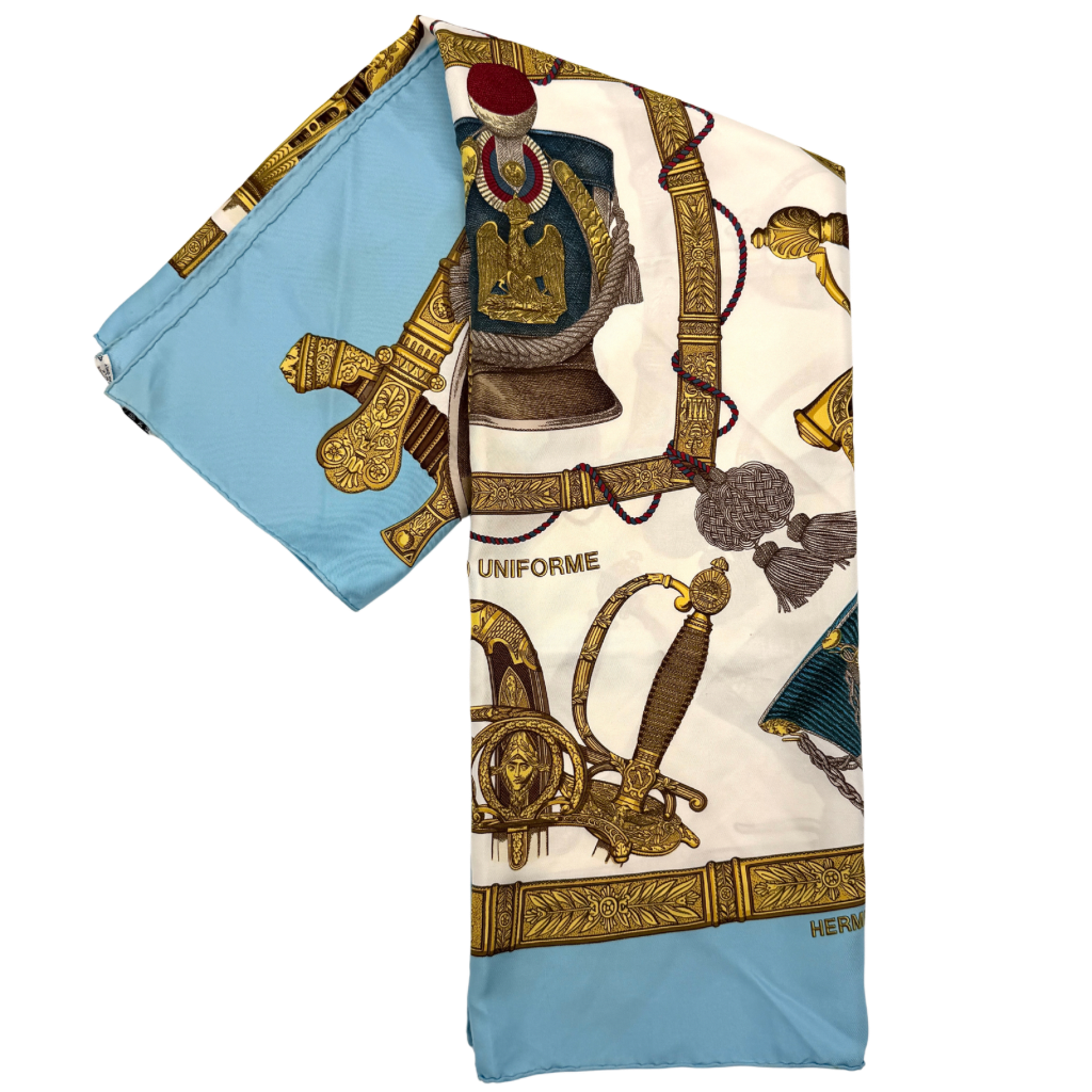 Hermès Grand Uniforme silk scarf with intricate gold and blue Napoleonic-inspired motifs framed on a soft blue and ivory background.