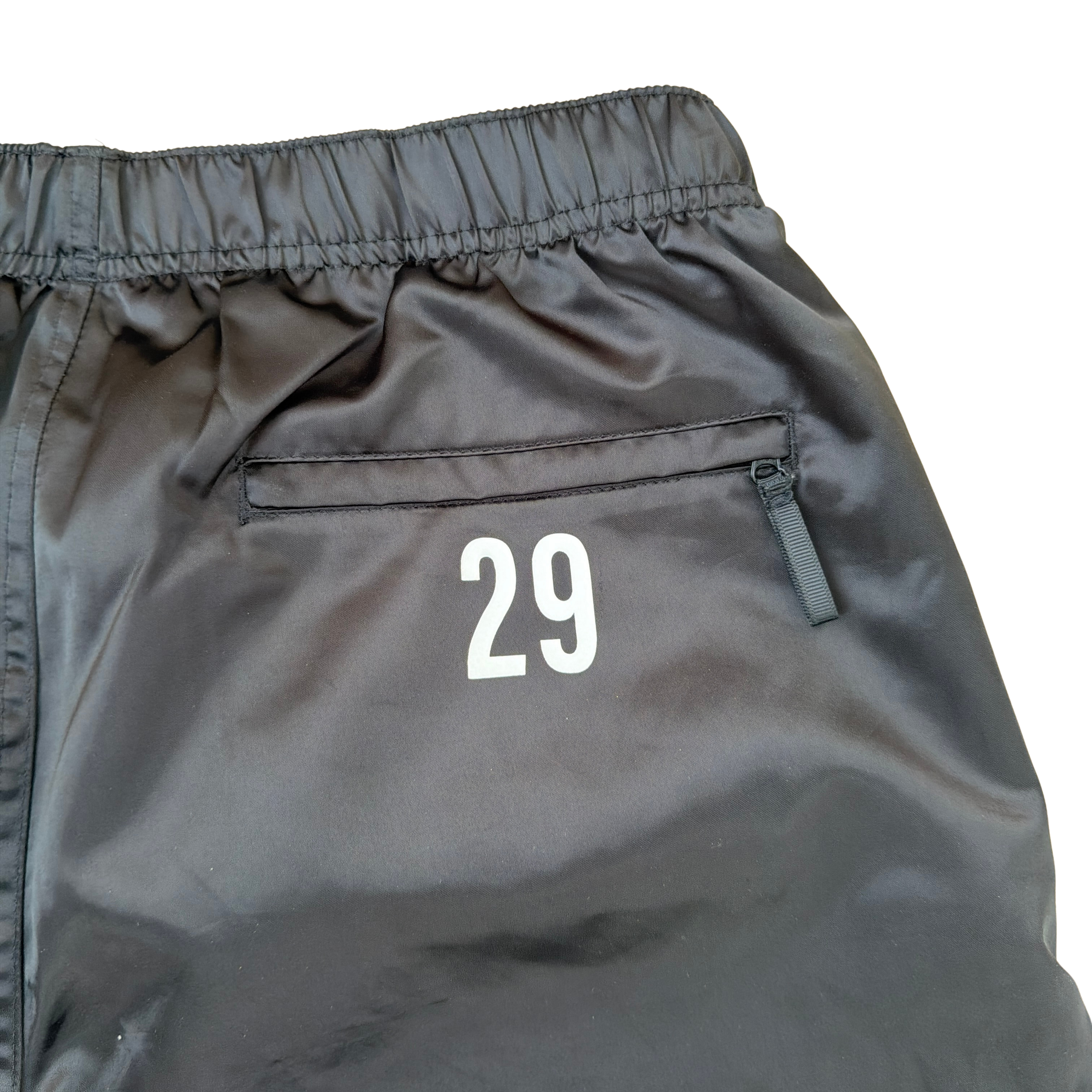 Supreme black satin track pants with white piping, a crest and Supreme logo embroidery at the hip, styled with relaxed legs.